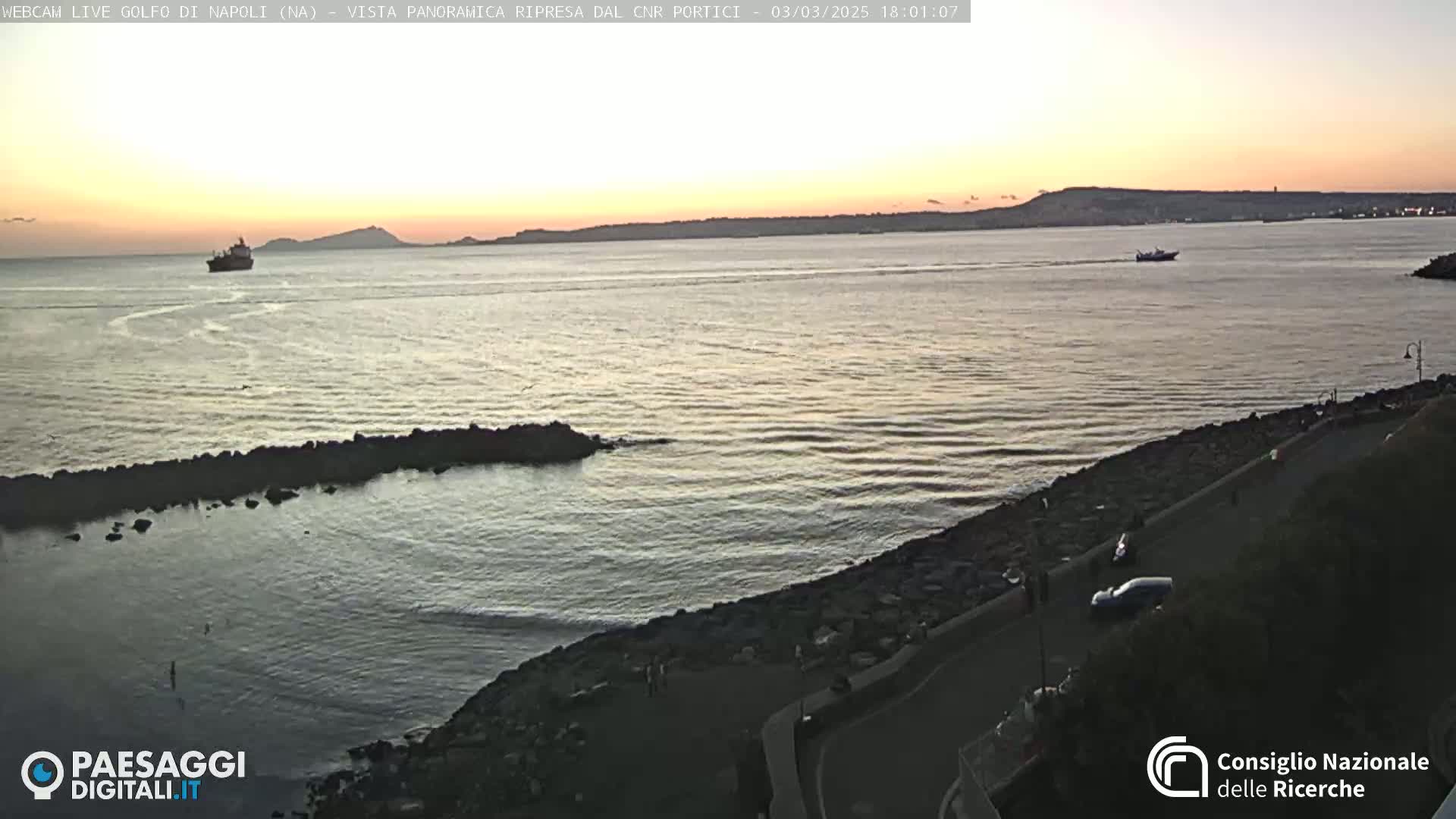 A calm sea at sunset with a few boats visible, a rocky shoreline, and a roadway with a few people present.