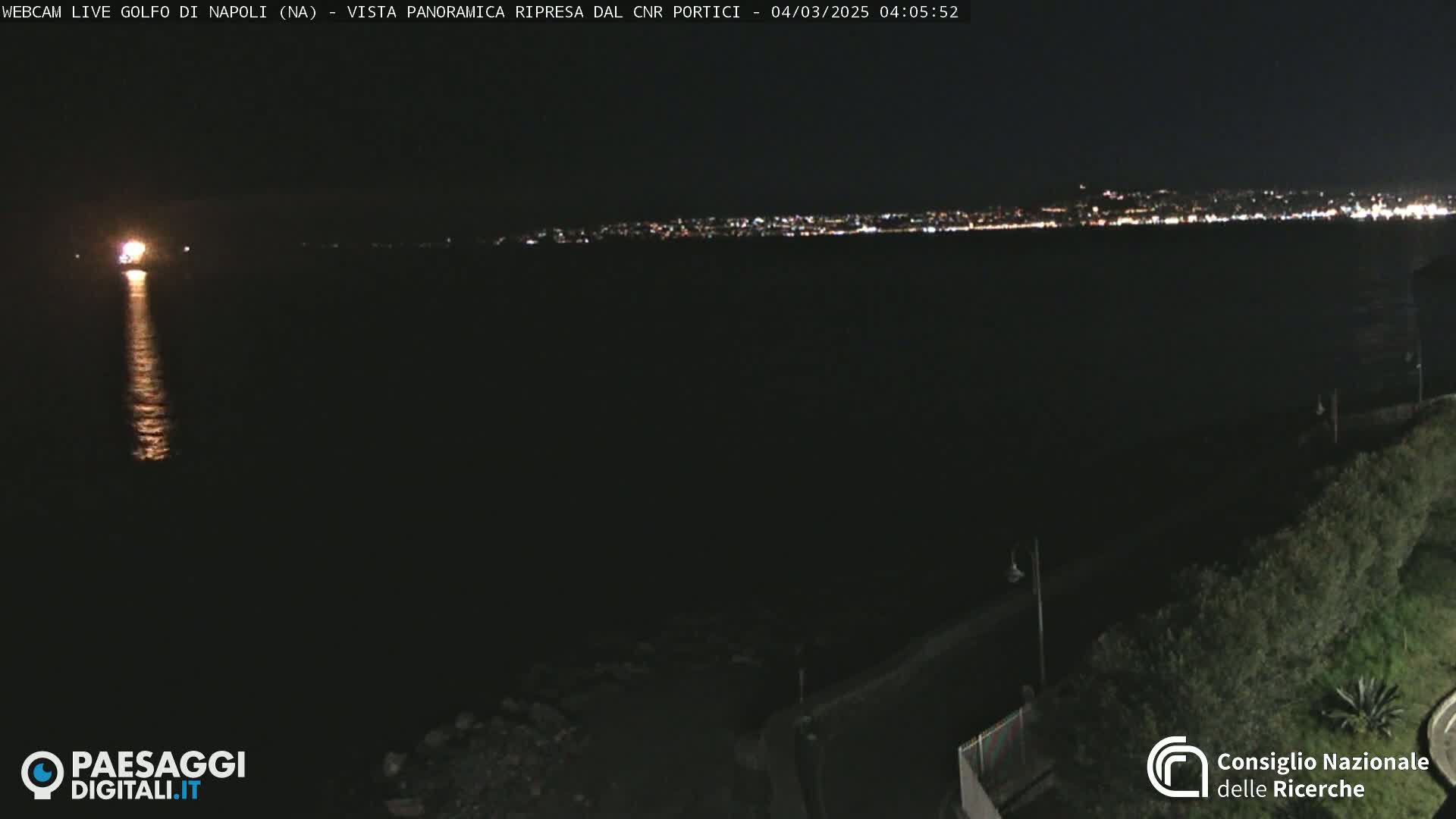 A nighttime coastal scene shows a dark body of water reflecting a distant bright light source and a city skyline illuminated by lights across the water.