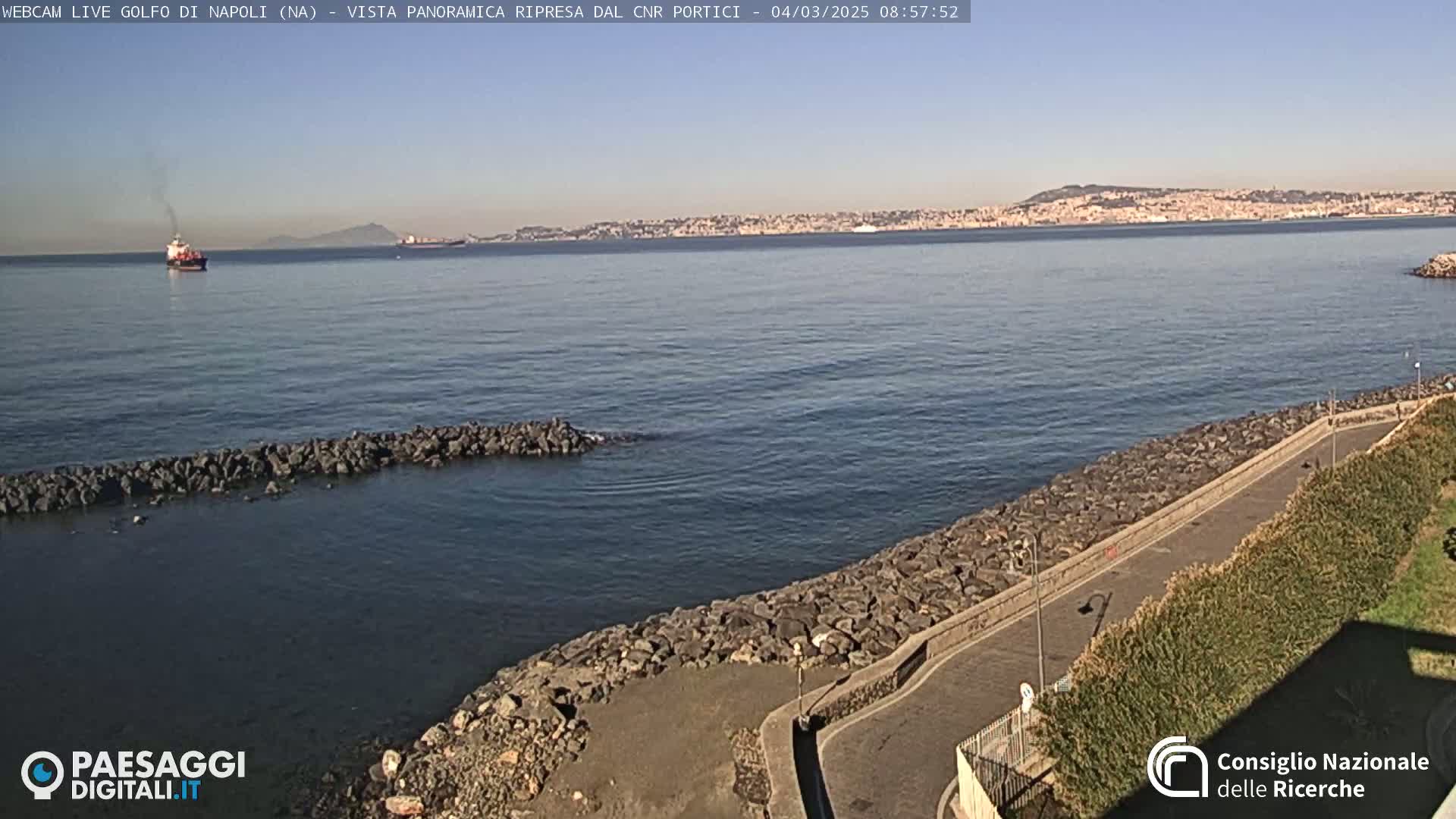 A rocky shoreline with a paved path curves alongside a calm sea, under a clear blue sky, with a distant city and a few ships visible on the horizon.