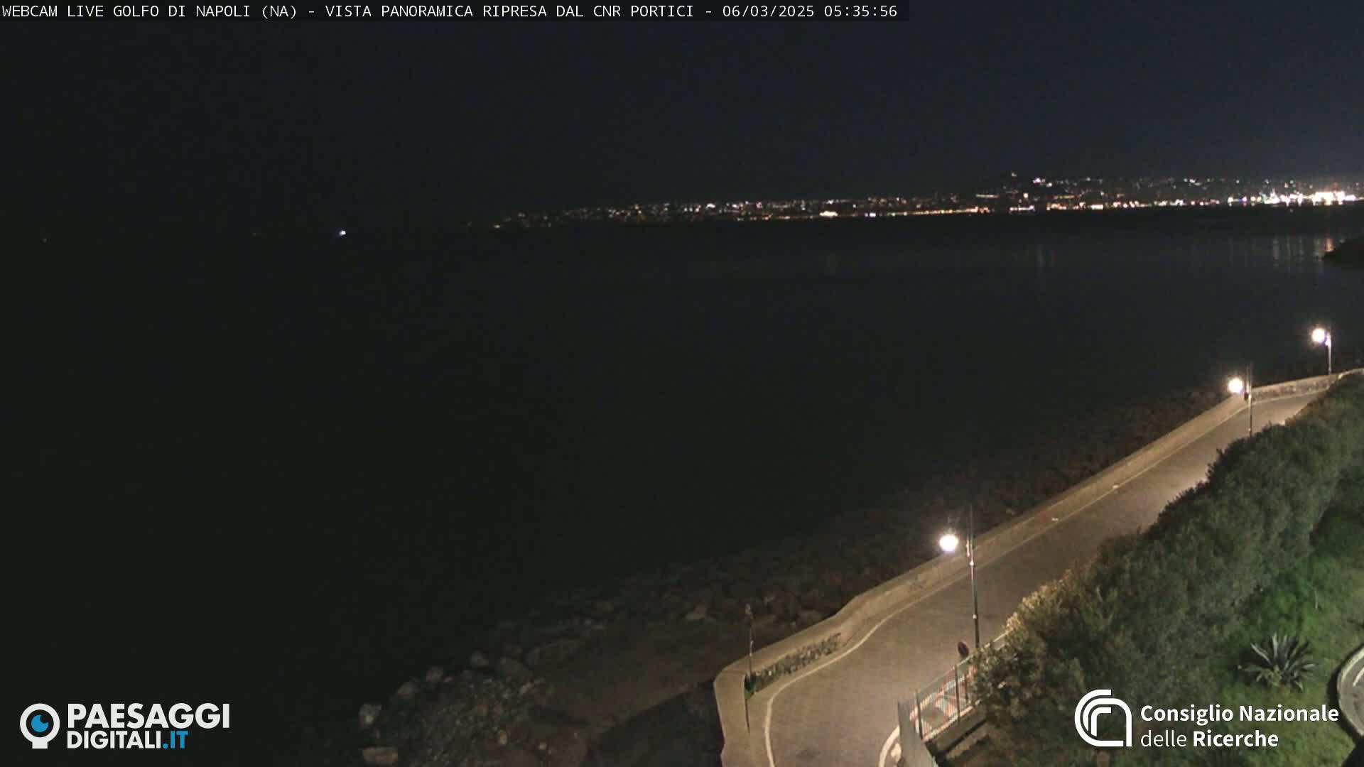 A nighttime, dark view of a paved road curving along a rocky shoreline, overlooking a dark body of water with a distant city skyline faintly illuminated.