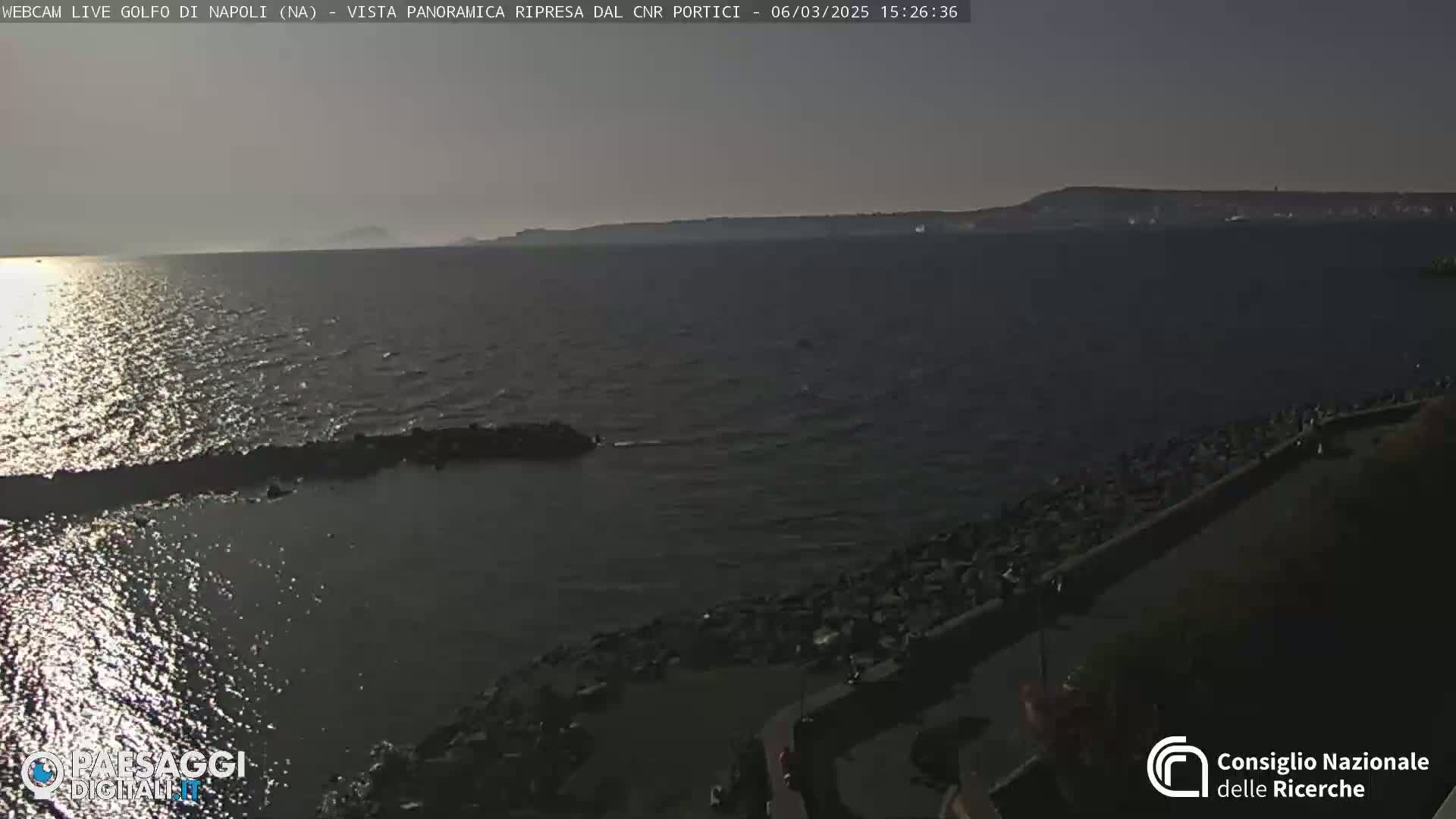 A calm, sunlit sea stretches to a distant shoreline under a clear sky, with a rocky embankment in the foreground.