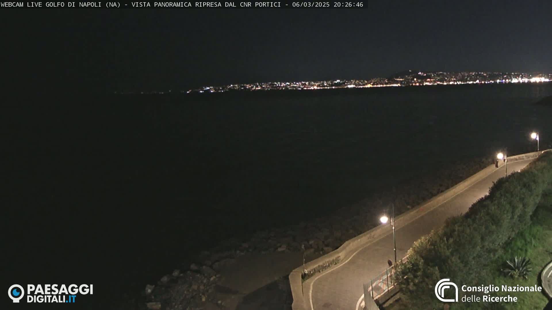 A nighttime, high-angle view shows a dark body of water with a city skyline faintly illuminated in the distance, and a paved road curving along a shoreline with vegetation and streetlights.