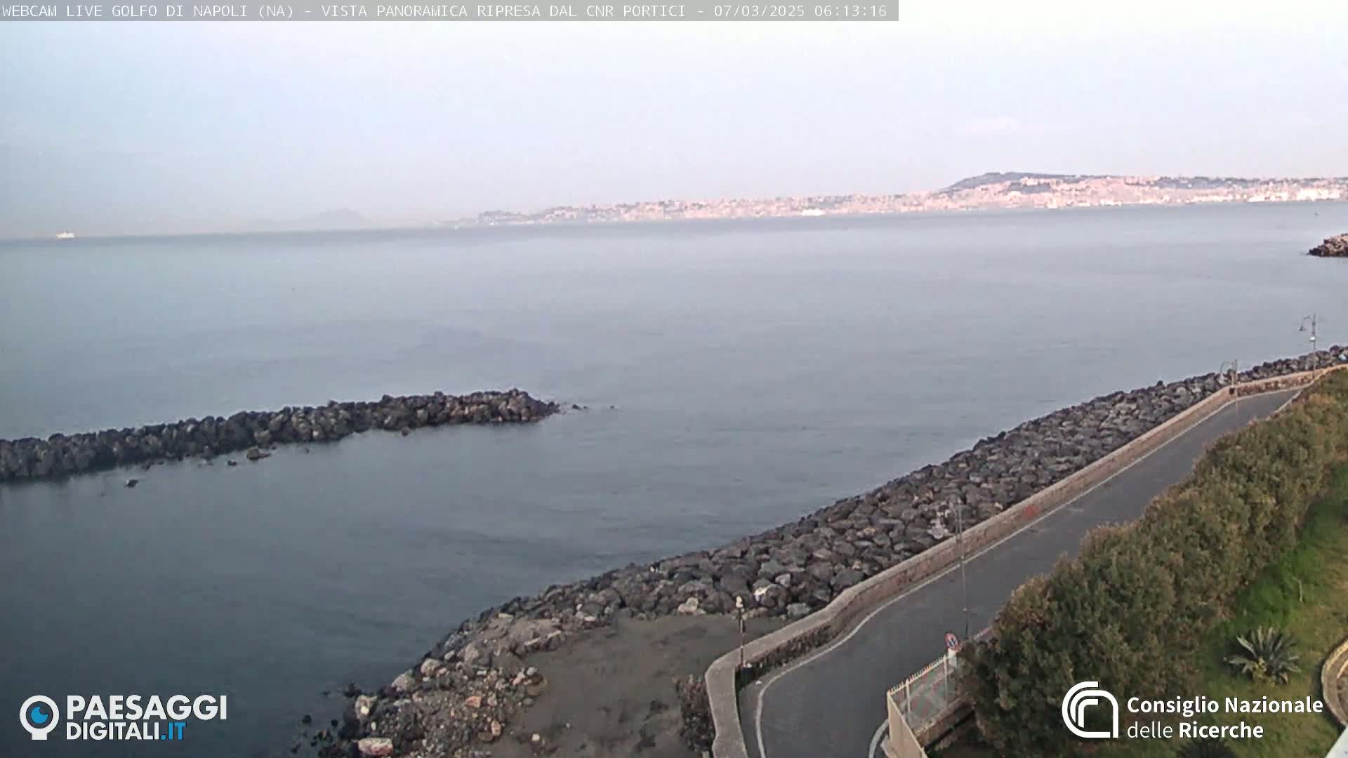 A rocky shoreline with a paved road curves alongside calm grey water, overlooking a distant city under a hazy sky.