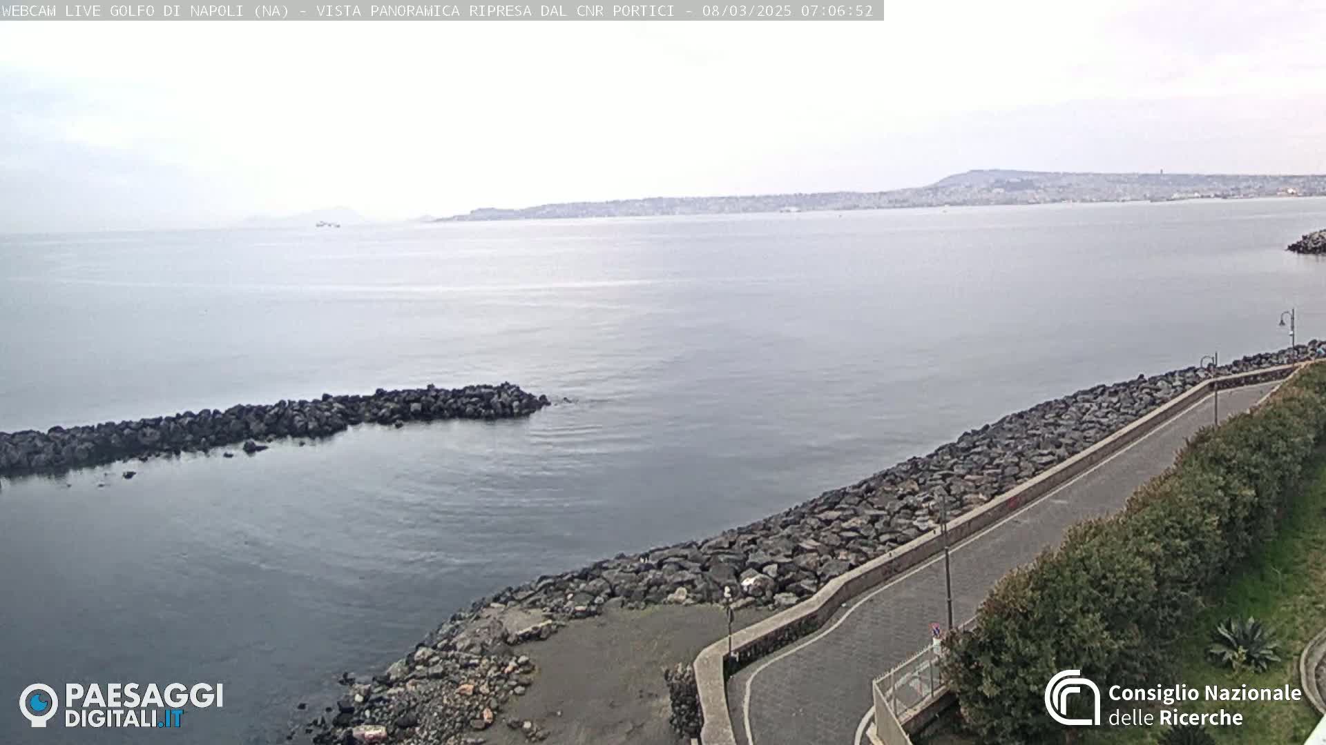 A rocky shoreline curves alongside a calm, gray body of water, with a distant city and hills visible under an overcast sky.