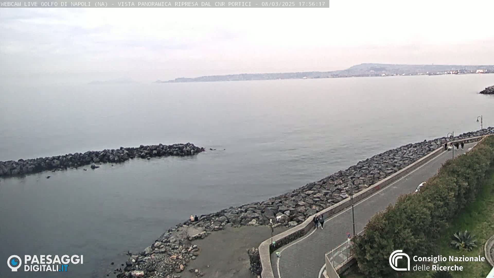 A calm, overcast day shows a rocky shoreline with a paved walkway beside a body of water extending to a hazy, distant coastline.