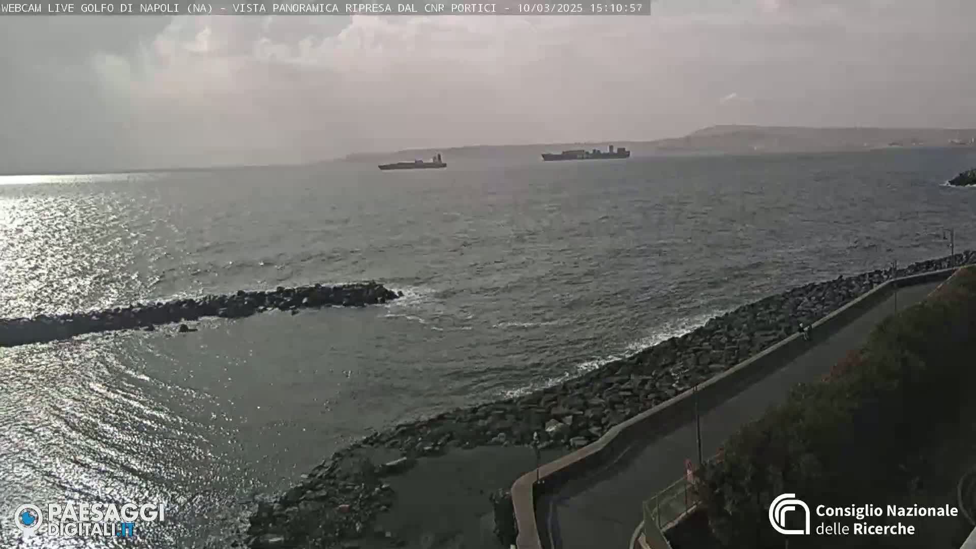 A partly cloudy day shows two cargo ships on the horizon, a rocky shoreline with a paved road, and some small waves.