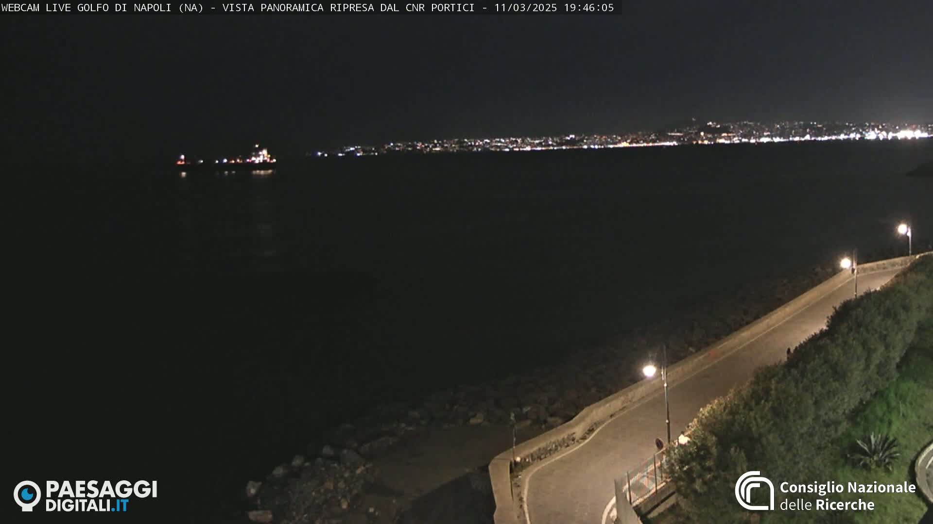 A nighttime, panoramic view of a dark body of water with a distant city skyline illuminated, a road curving along a rocky shoreline, and a single vessel on the water.