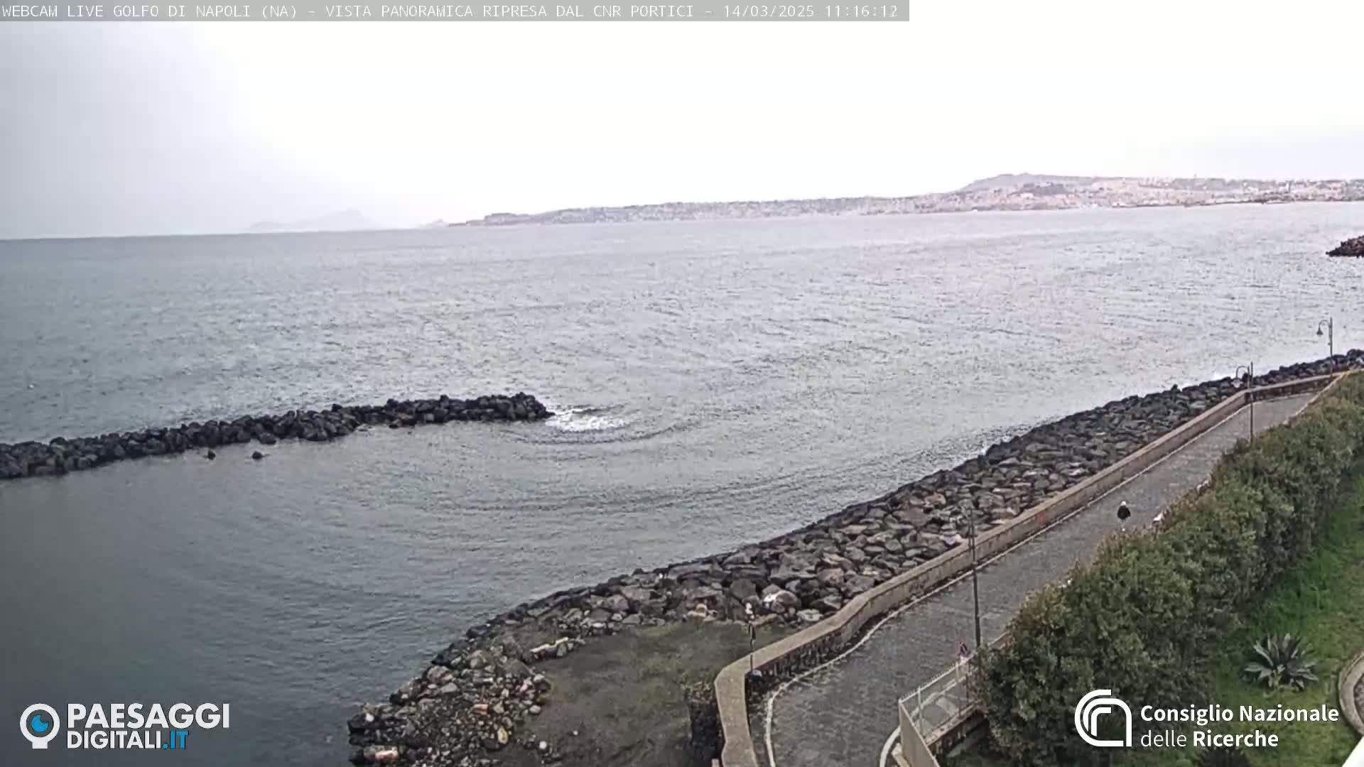 A rocky shoreline with a paved path curves alongside calm, gray water under an overcast sky, with a distant landmass visible on the horizon.