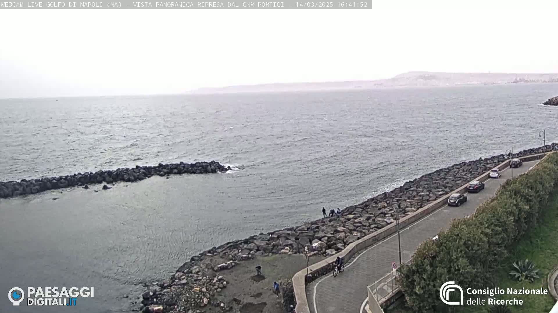 A rocky coastline curves alongside a calm, gray sea under an overcast sky, with several cars parked along a road above the shore.