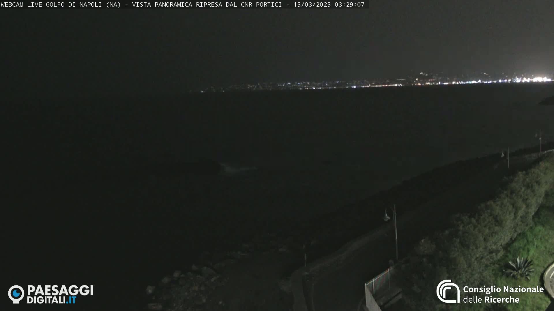 A nighttime, panoramic view of a dark body of water with a distant, faintly lit cityscape under a clear sky.