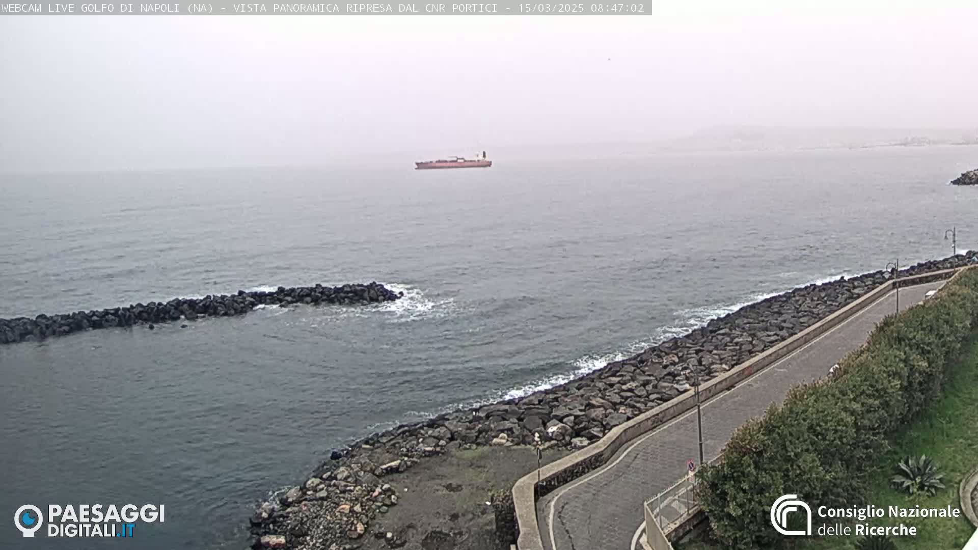 A hazy view of a calm sea with a cargo ship in the distance, alongside a rocky shoreline with a road and vegetation.