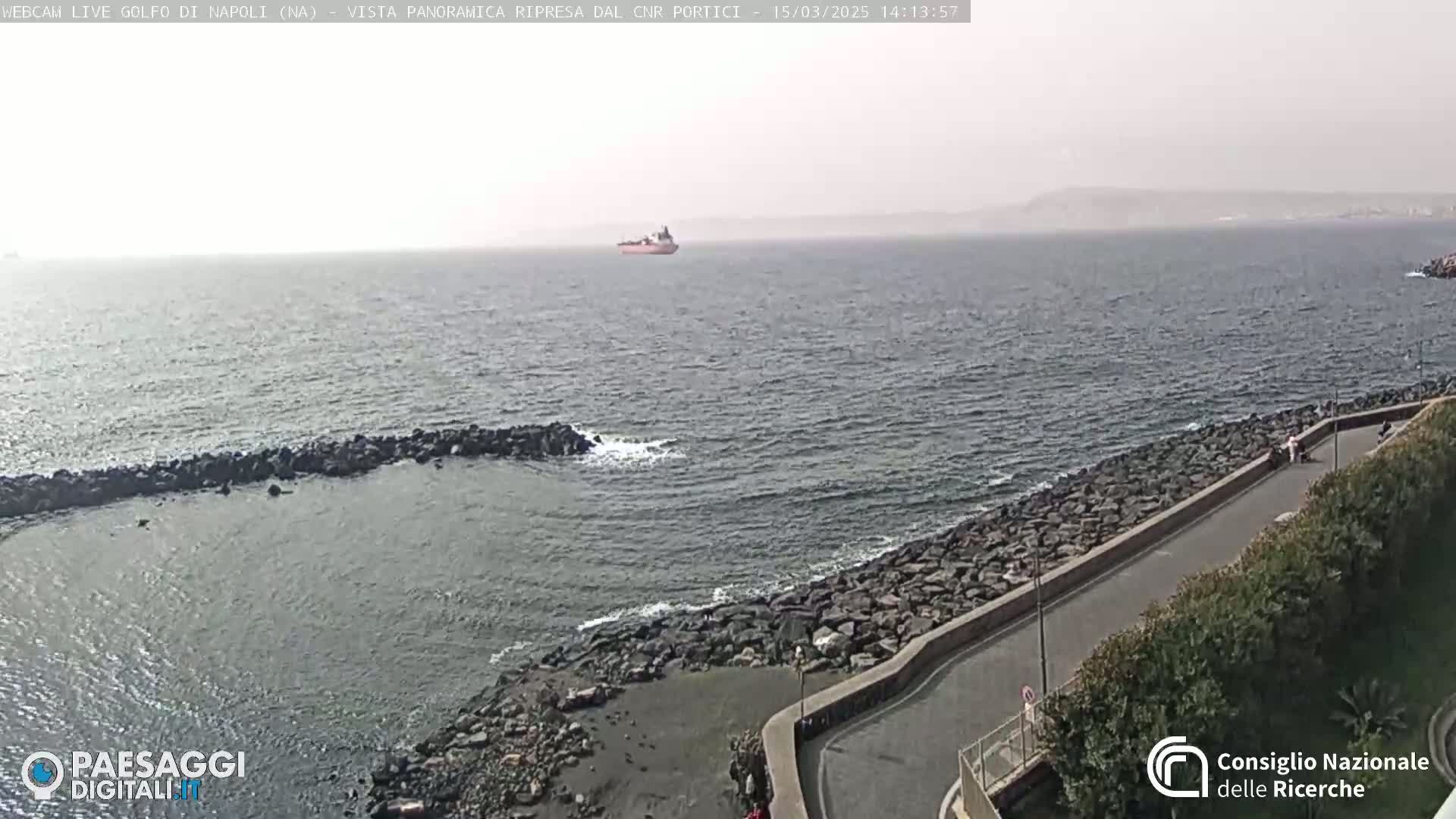 A hazy, distant view of a large ship on the water, seen from a rocky coast with a paved road curving along the shoreline.