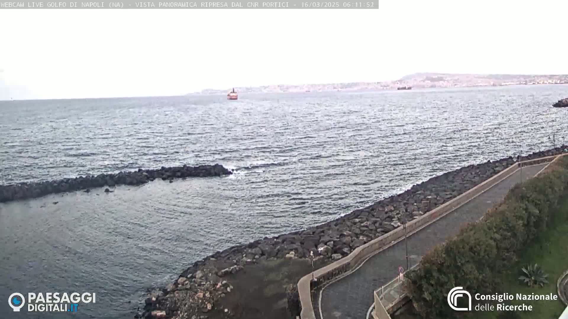 A rocky shoreline with a paved path curves gently around a calm, gray-blue sea under an overcast sky, a distant cityscape and a lone vessel visible on the horizon.