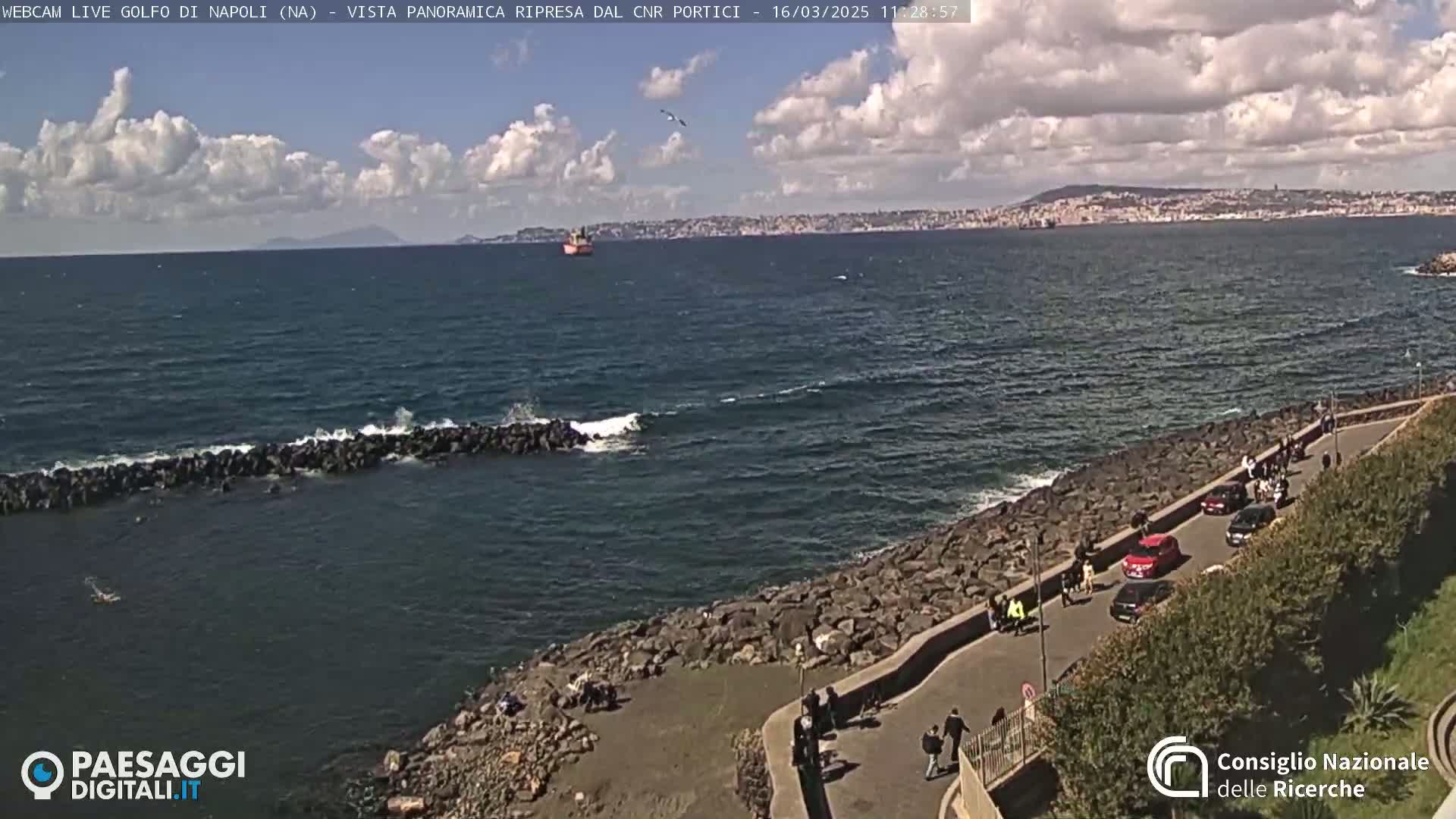 A rocky coastline with a paved walkway and parked cars overlooks a partly cloudy sea with small waves breaking against a breakwater; a distant city and mountains are visible across the water.