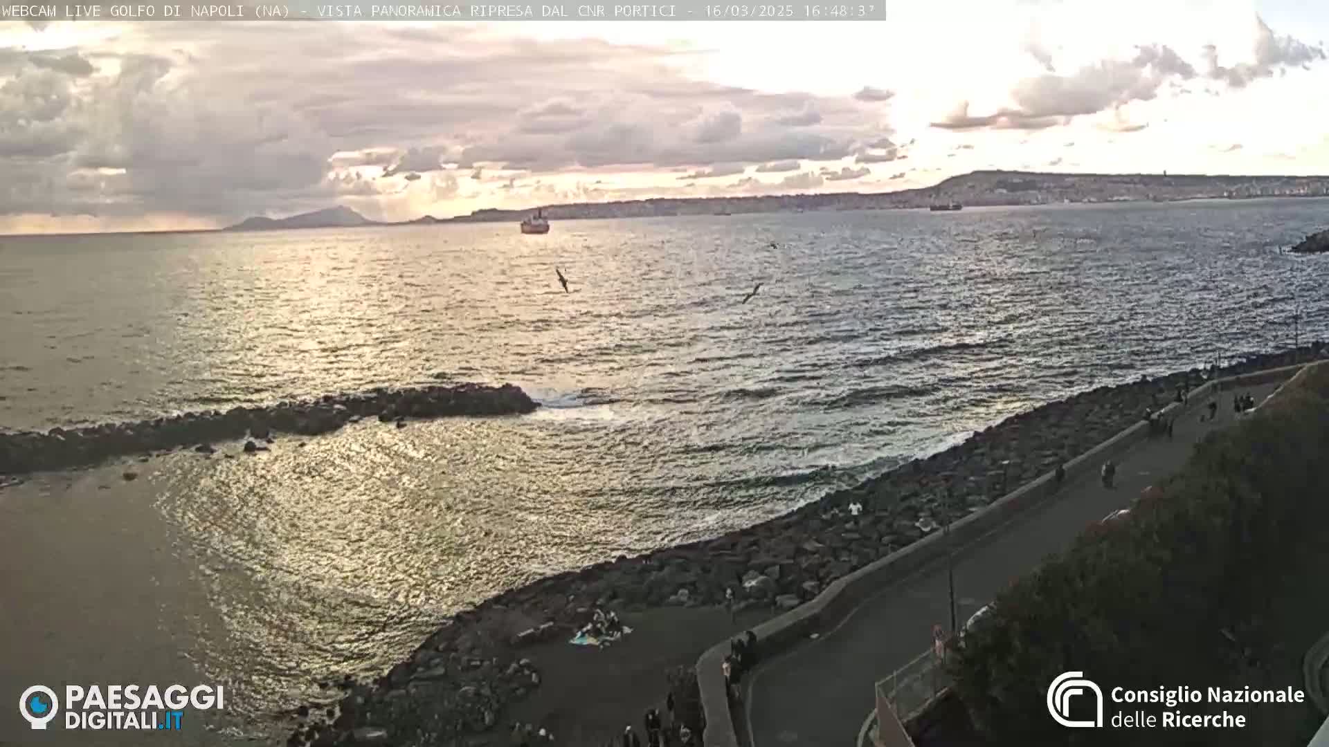 A rocky coastline with a paved road overlooks a calm sea under a partly cloudy sunset sky.