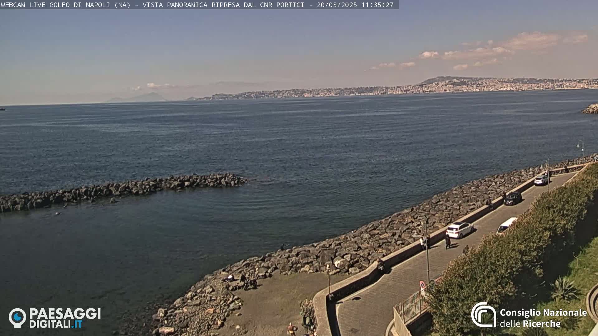 A coastal view shows a rocky shoreline with a road and parked cars, overlooking a calm body of water under a partly cloudy sky, with a distant city visible in the background.