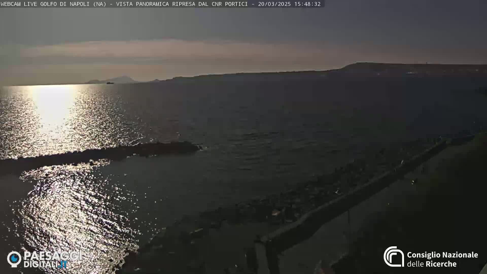 A calm sea reflects the setting sun, with a distant shoreline visible under a mostly clear sky.