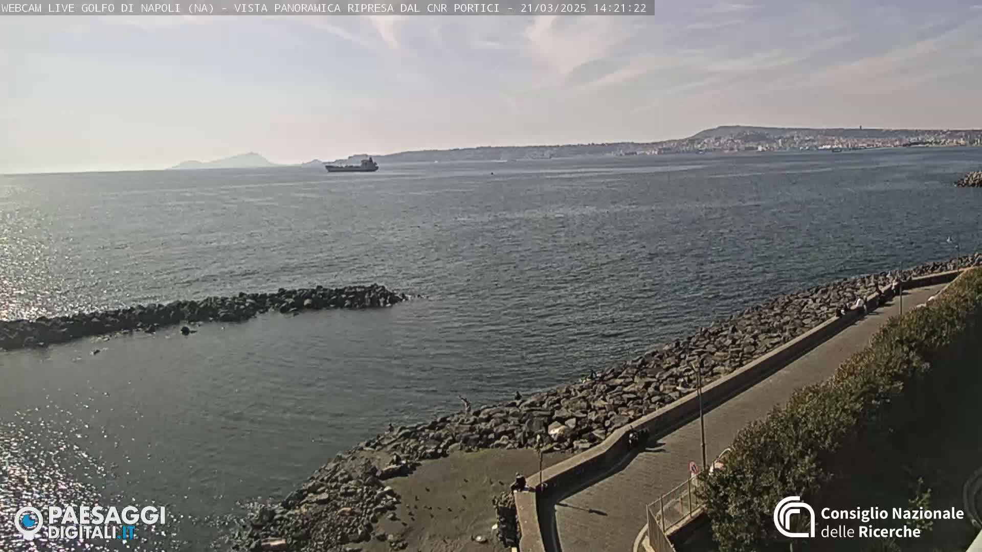 A mostly sunny day overlooking a calm body of water with a rocky shoreline and a paved walkway, a distant cityscape, and a large ship visible on the horizon.