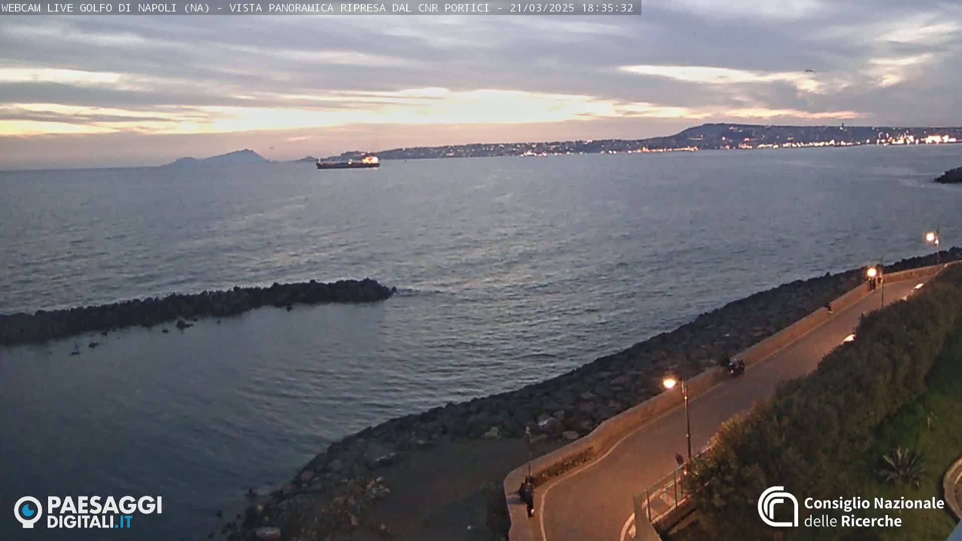 A twilight view of a calm sea with a rocky shore and a road curving along the coastline, with a distant city skyline illuminated at night.