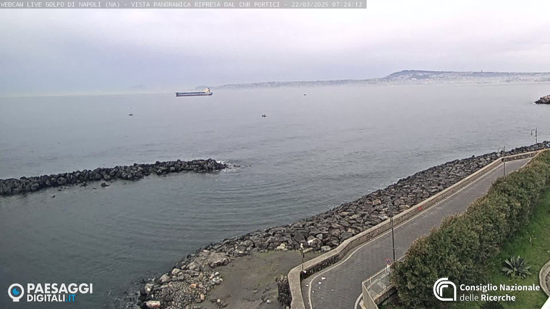 A mostly cloudy day shows a calm sea with a large cargo ship and a distant city skyline, seen from a rocky shoreline with a paved road curving along it.