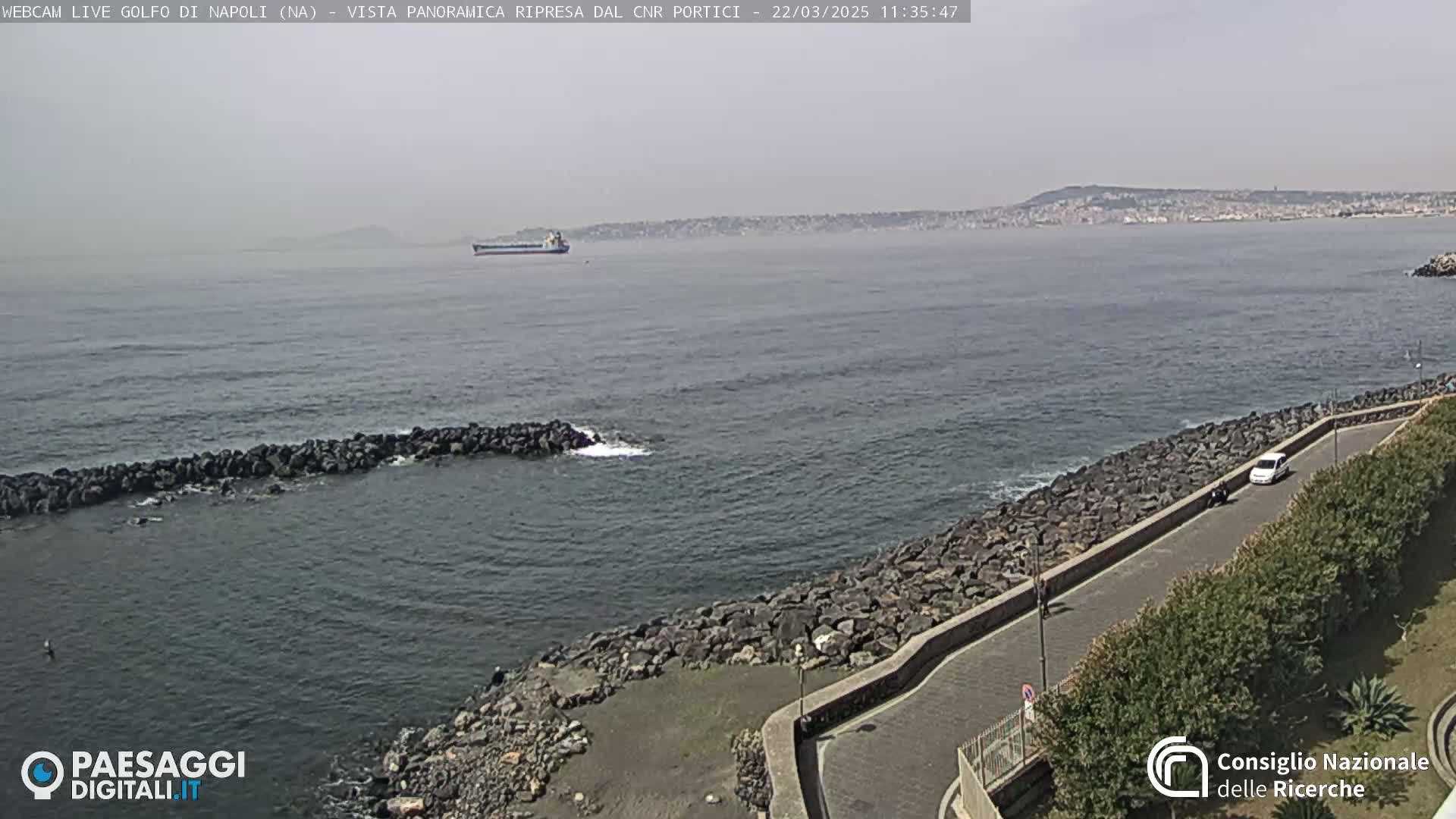 A hazy, daytime view of a calm sea with a large cargo ship in the distance, a rocky shoreline with a road, and sparse vegetation.