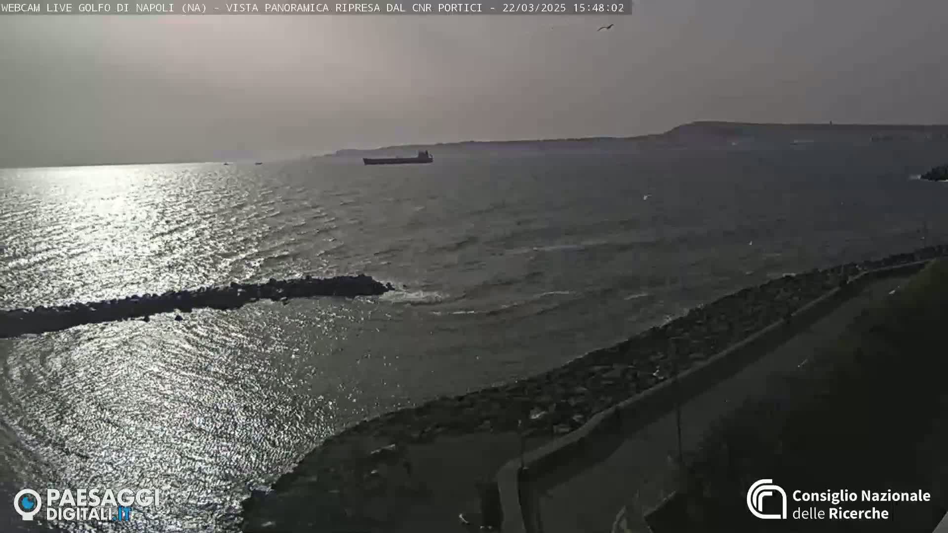 A slightly overcast day shows a calm sea with a large cargo ship and a rocky shoreline.