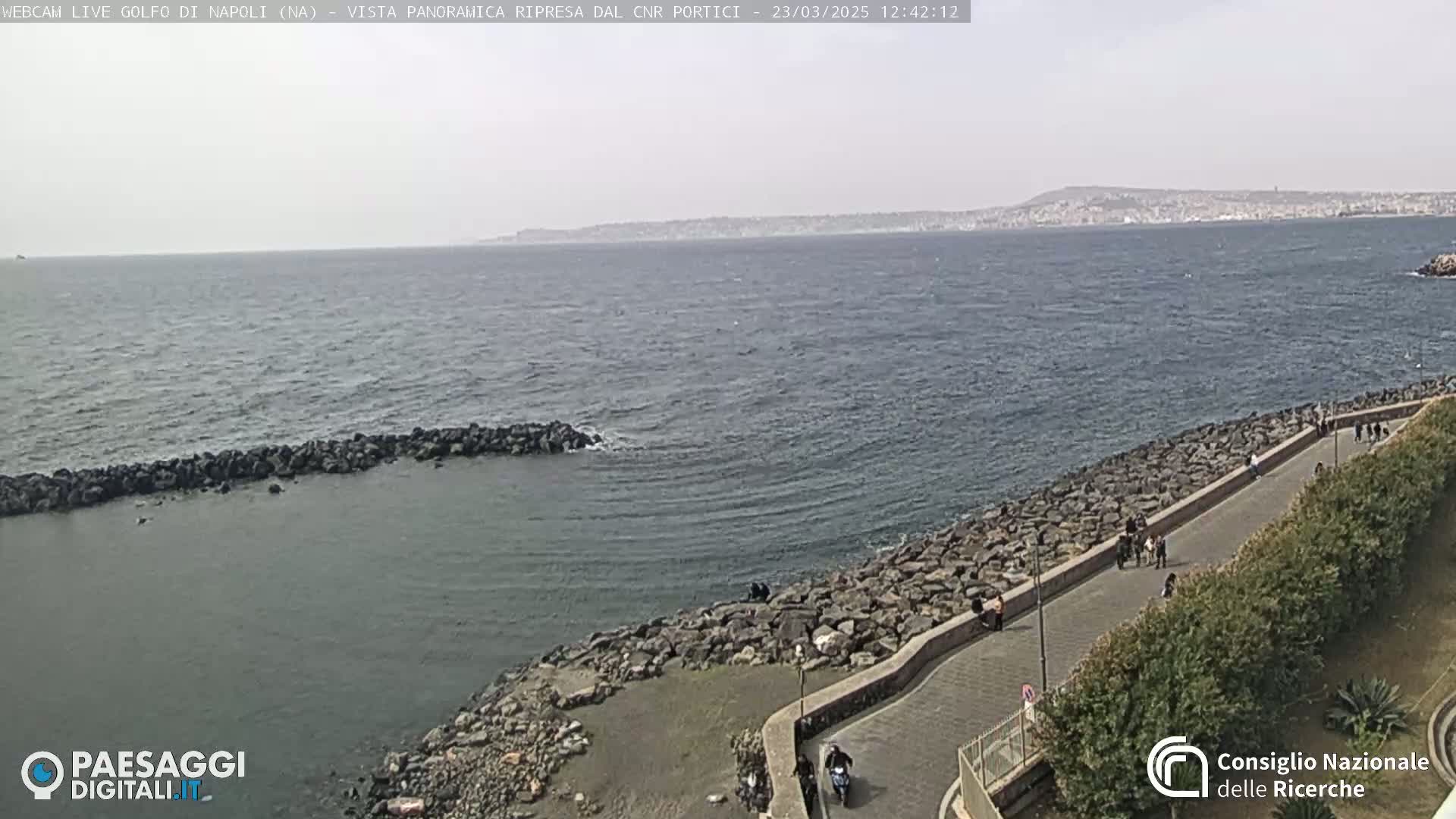 A rocky shoreline with a paved walkway and sparse vegetation overlooks a calm sea under a hazy sky, with a distant city visible across the water.