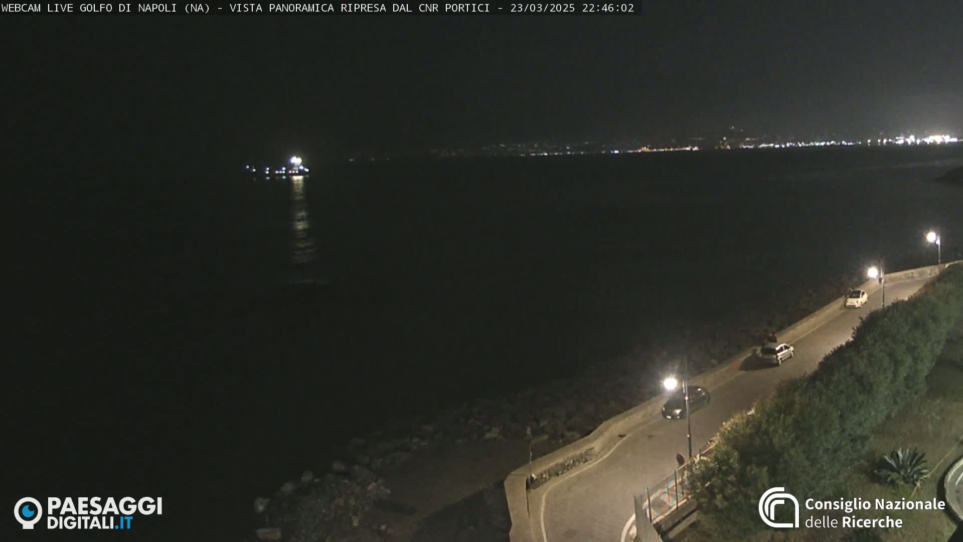 A nighttime view of a dark body of water with a distant, faintly lit shoreline, and a road with a few cars visible along the coast.