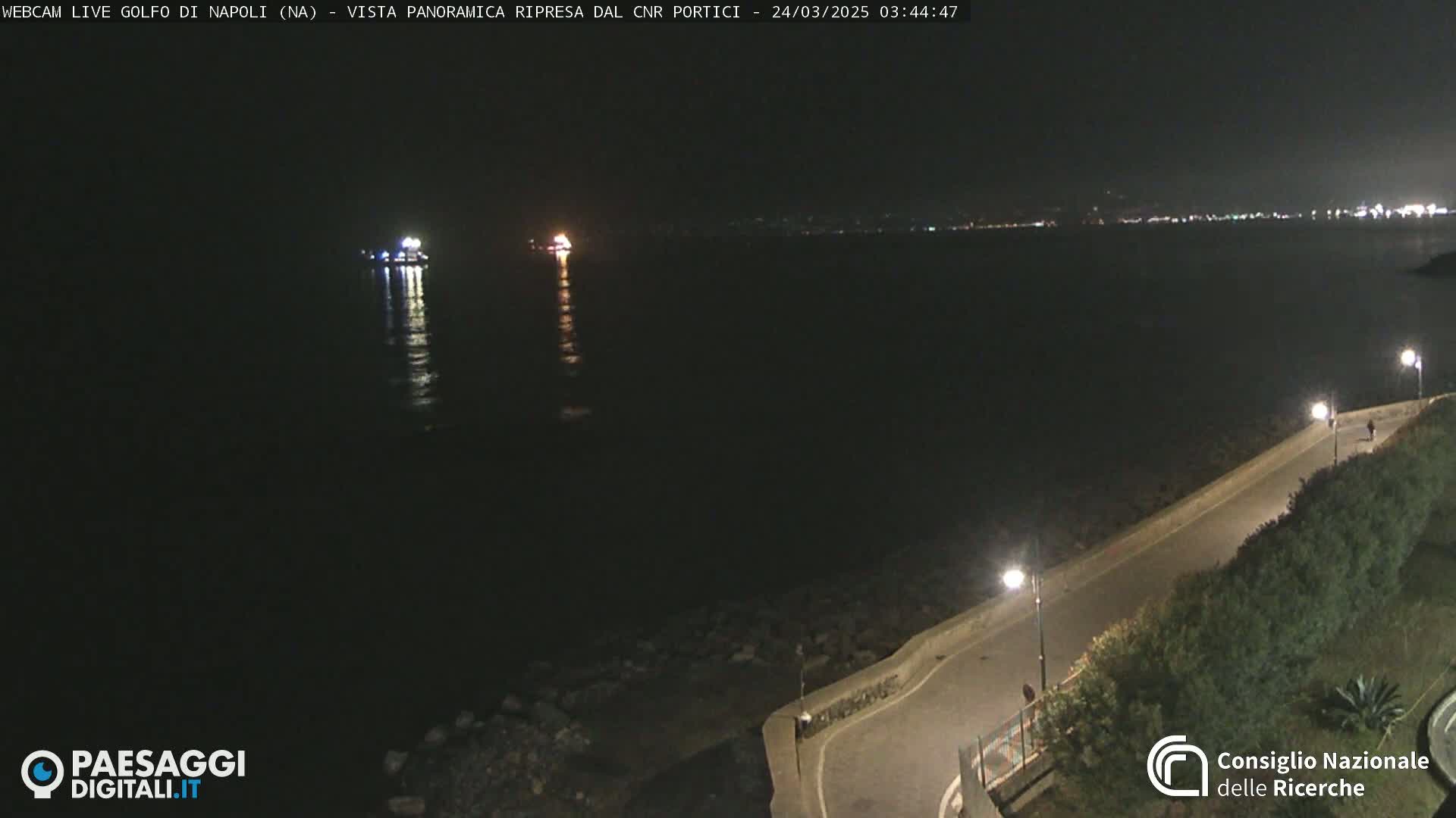 A nighttime view of a dark body of water with a distant light source and a paved walkway with streetlights along a shoreline.