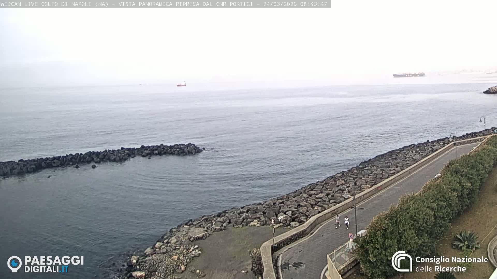 A mostly overcast view of a rocky coastline and calm sea with a few small boats in the distance, and two people walking along a paved path next to a low wall.