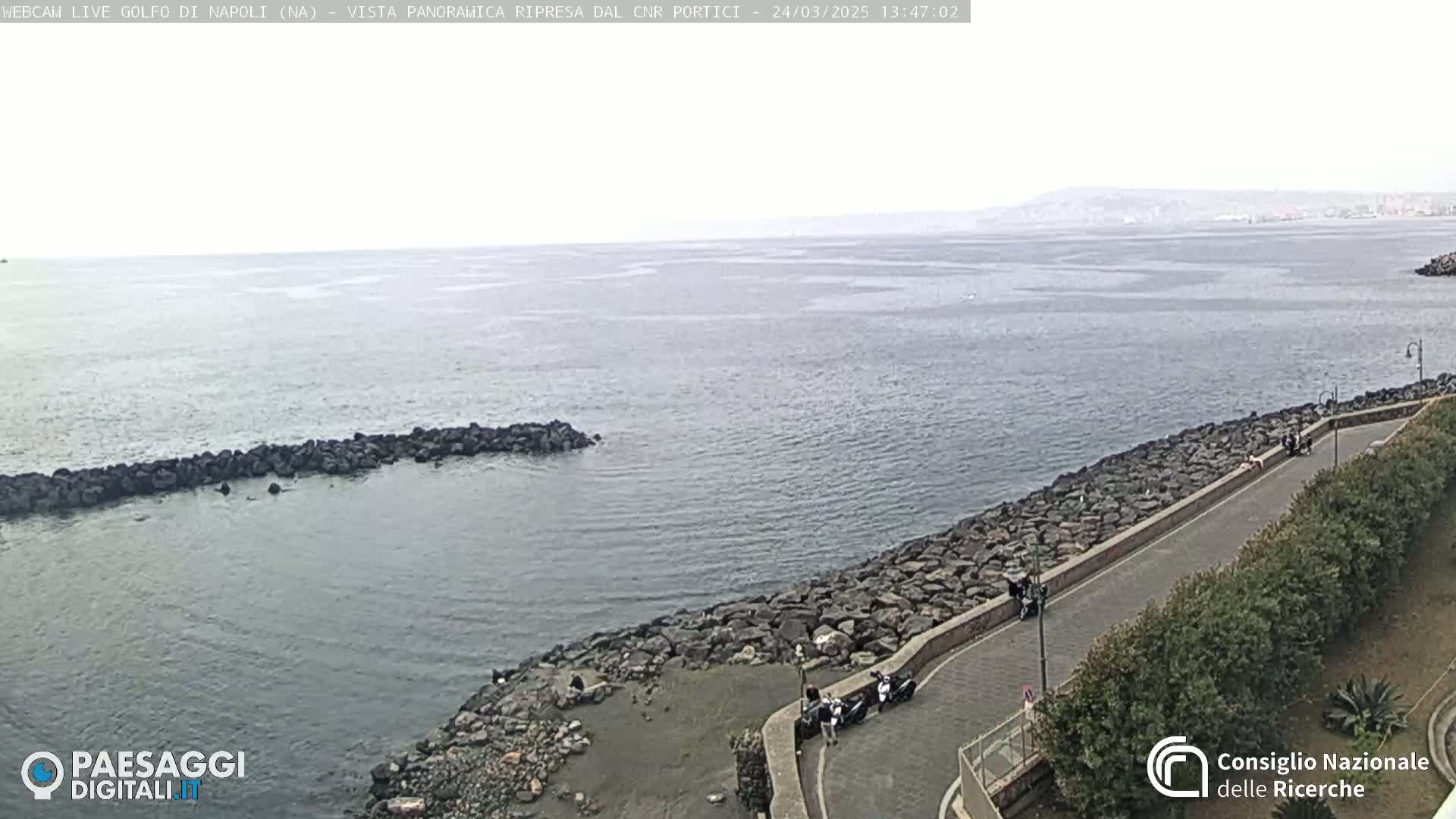 A rocky shoreline curves alongside a calm sea under an overcast sky, with a distant landmass visible on the horizon.