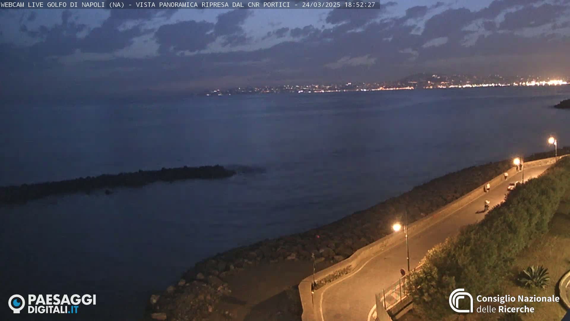 A nighttime, coastal scene shows a dark-blue sea under a partly cloudy sky, with a roadway curving along a rocky shoreline, and distant city lights visible across the water.