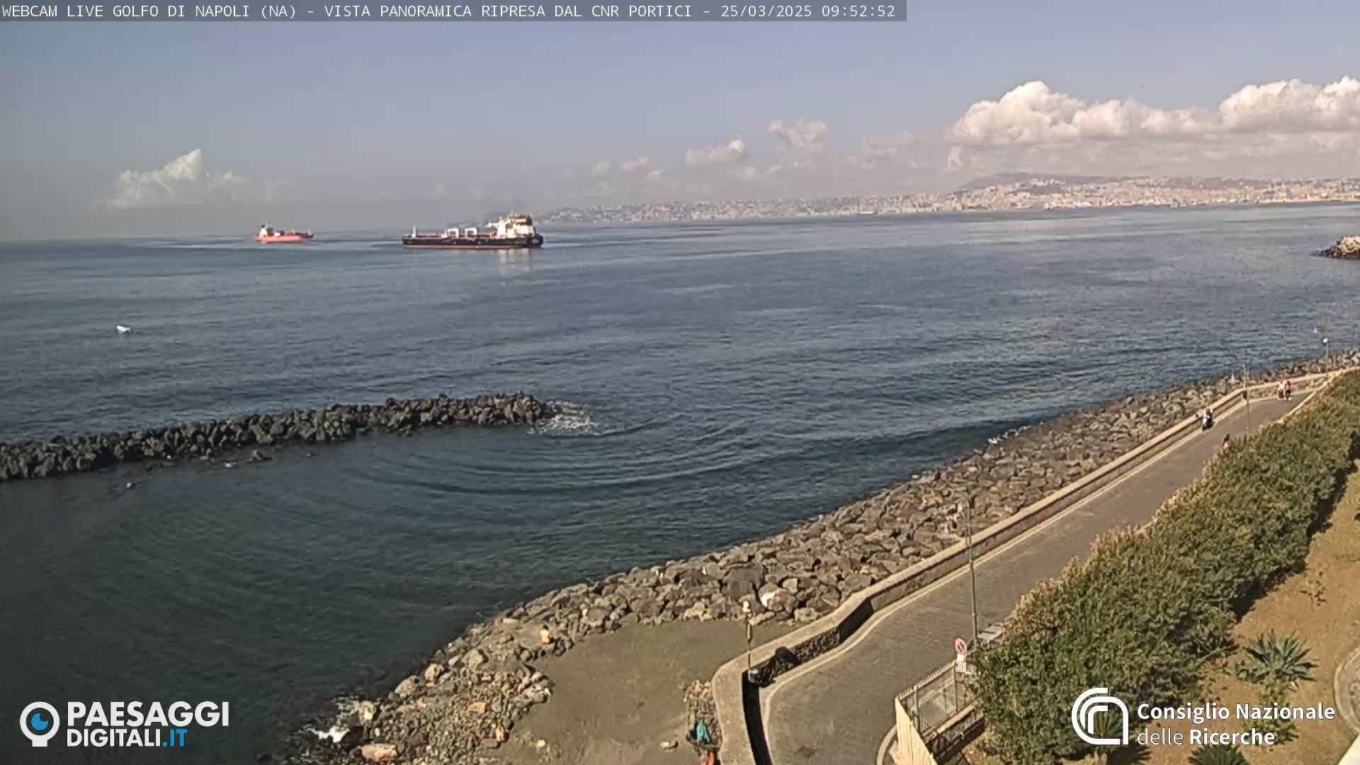 A partly cloudy day shows a coastal road next to a rocky shoreline, with two ships visible in the distance across a calm sea.