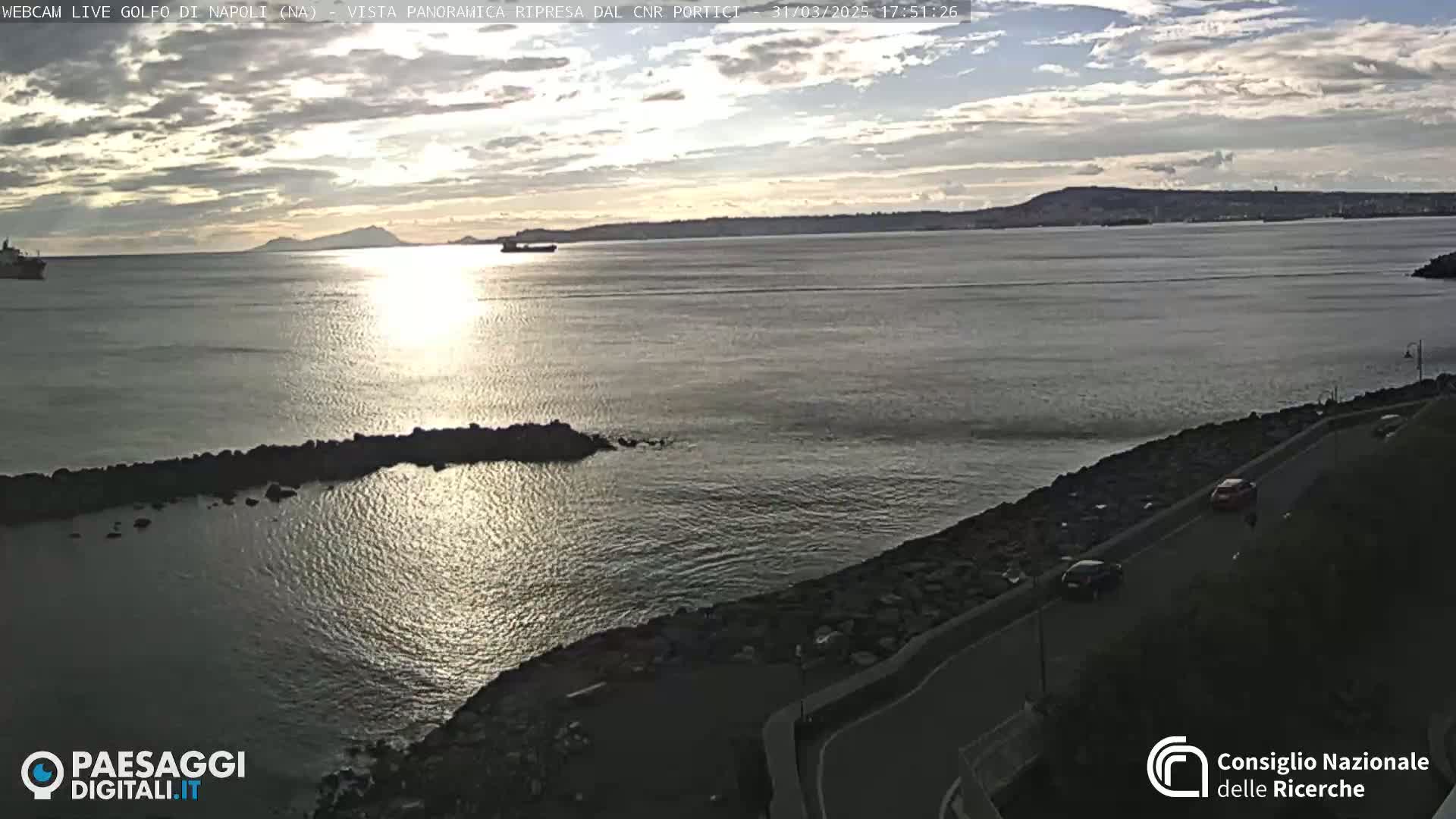 A calm, sunlit body of water stretches to a distant shoreline under a partly cloudy sky, with a road and some cars visible in the foreground.