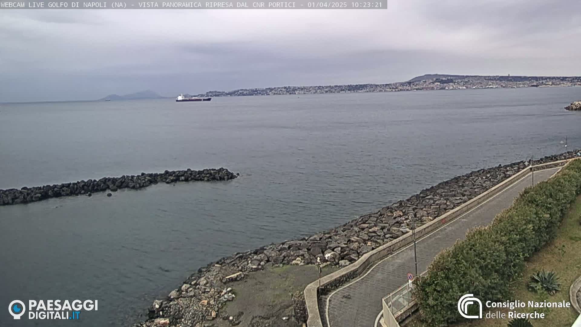 A coastal road curves along a rocky shoreline overlooking a calm bay and a distant city under a cloudy sky.