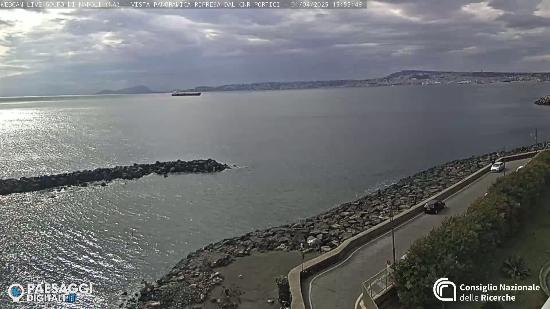 A partly cloudy day reveals a panoramic view of a calm sea with a rocky shoreline and a road curving along the coast, featuring a few cars.