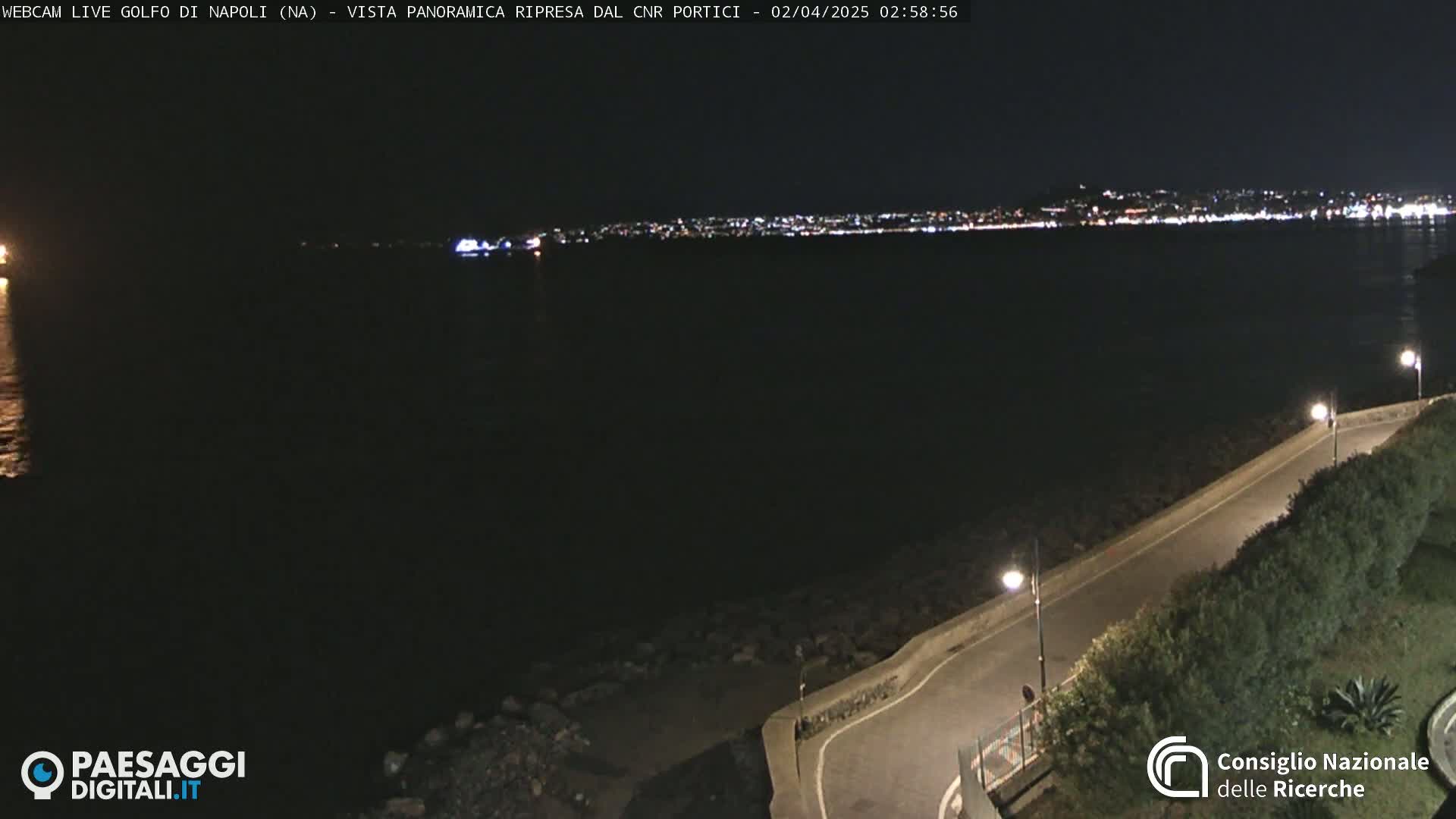 A nighttime view of a dark body of water with a city's lights visible in the distance across the water, and a dark roadway curving along the water's edge, lit by streetlights.