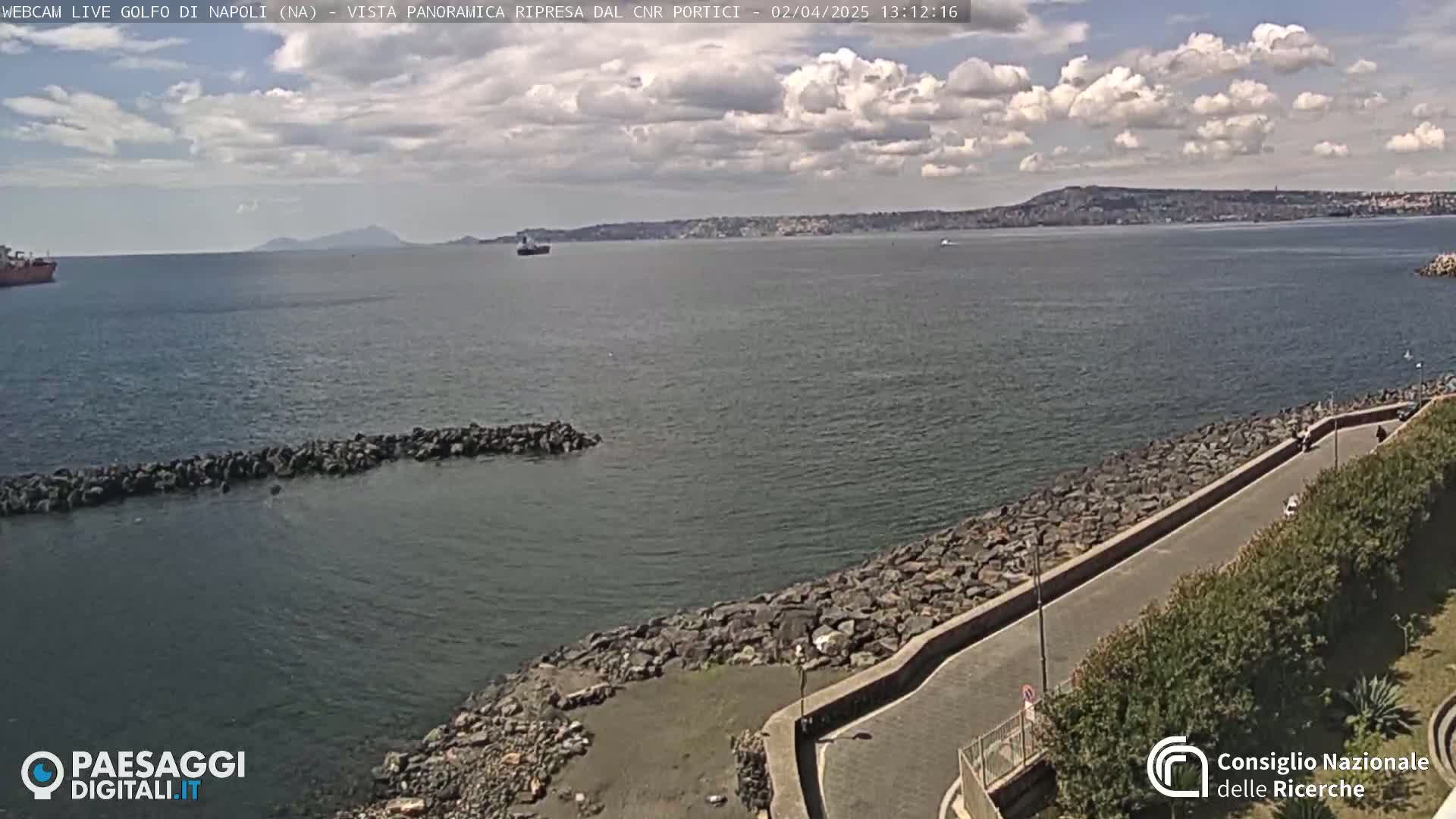 A partly cloudy day reveals a wide expanse of calm sea, a rocky shoreline with a paved walkway, and distant land in the background.