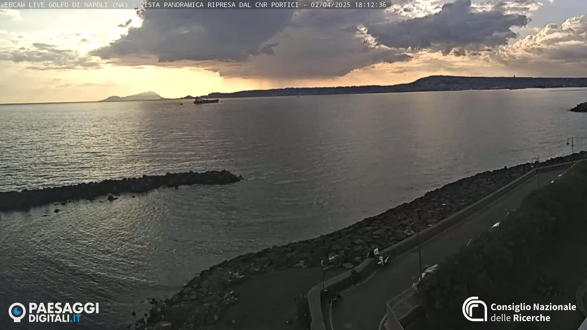 A calm sea under a partly cloudy sunset sky, viewed from a rocky shore with a road winding along the coast.