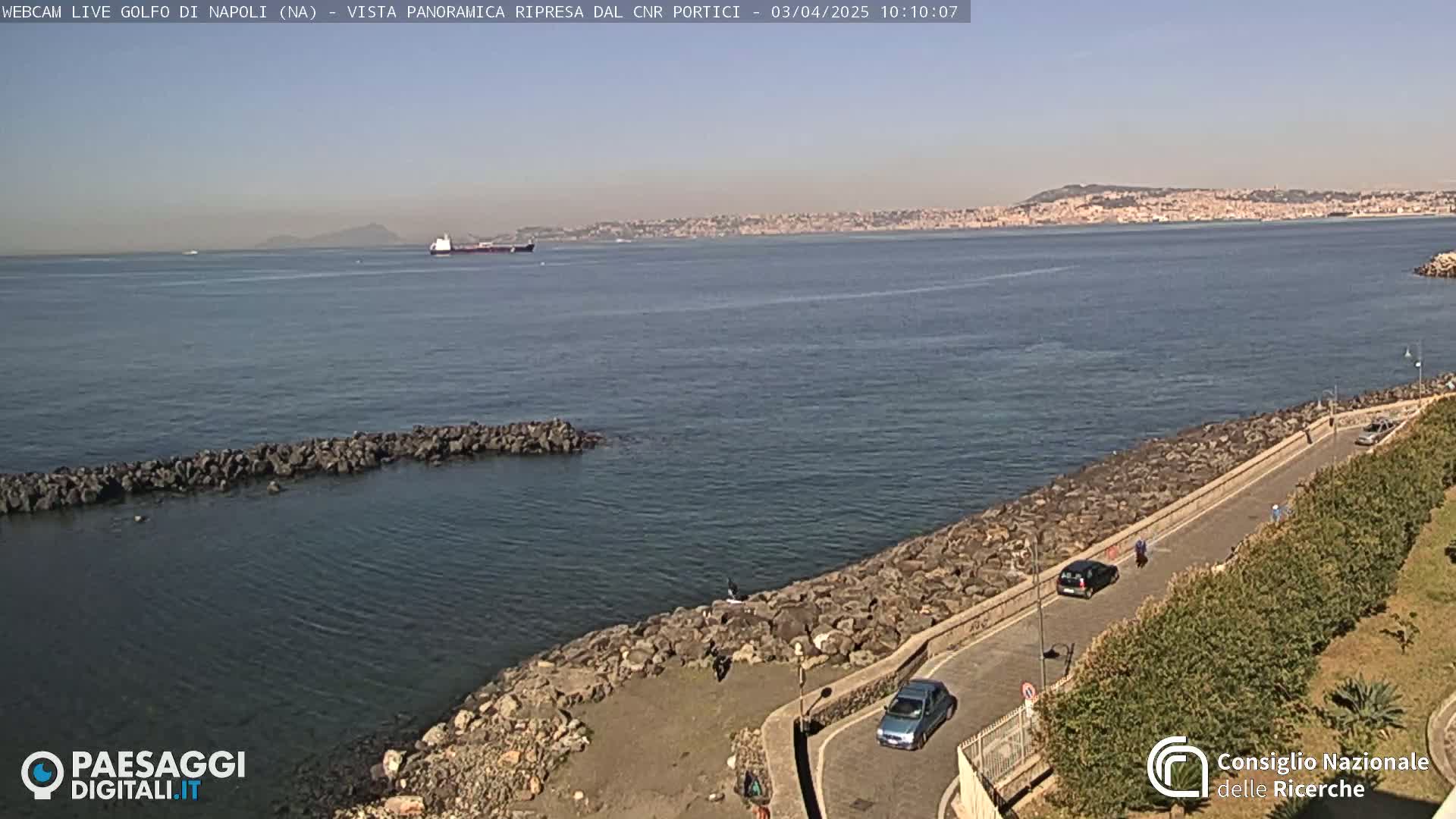 A calm, sunny day shows a rocky shoreline with a road and a few cars, overlooking a bay with a large cargo ship and a distant cityscape under a clear sky.