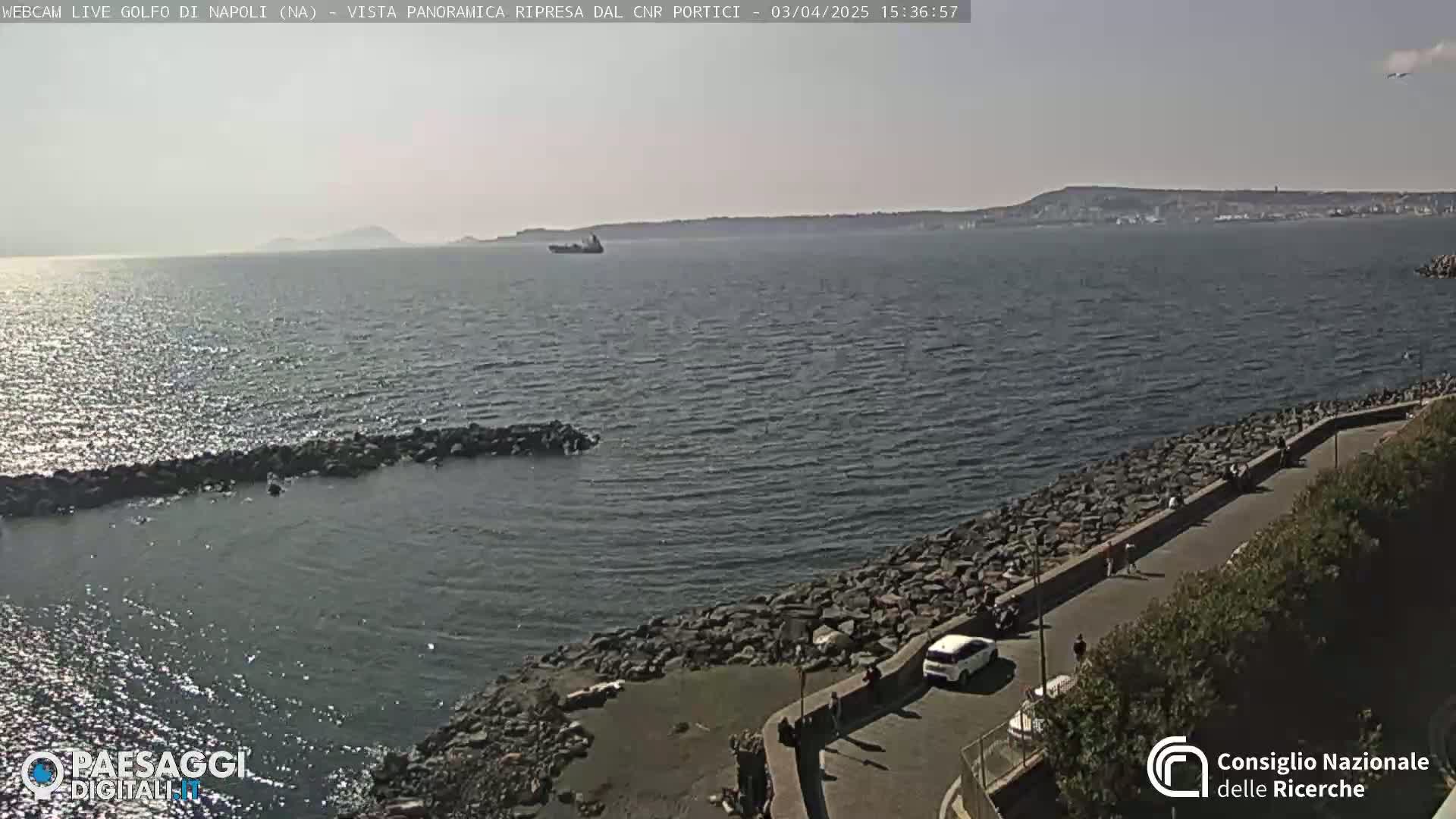 A calm, sunny day overlooks a rocky shoreline with a few people walking along a path next to the water; a distant coastline and a large ship are visible across the bay.