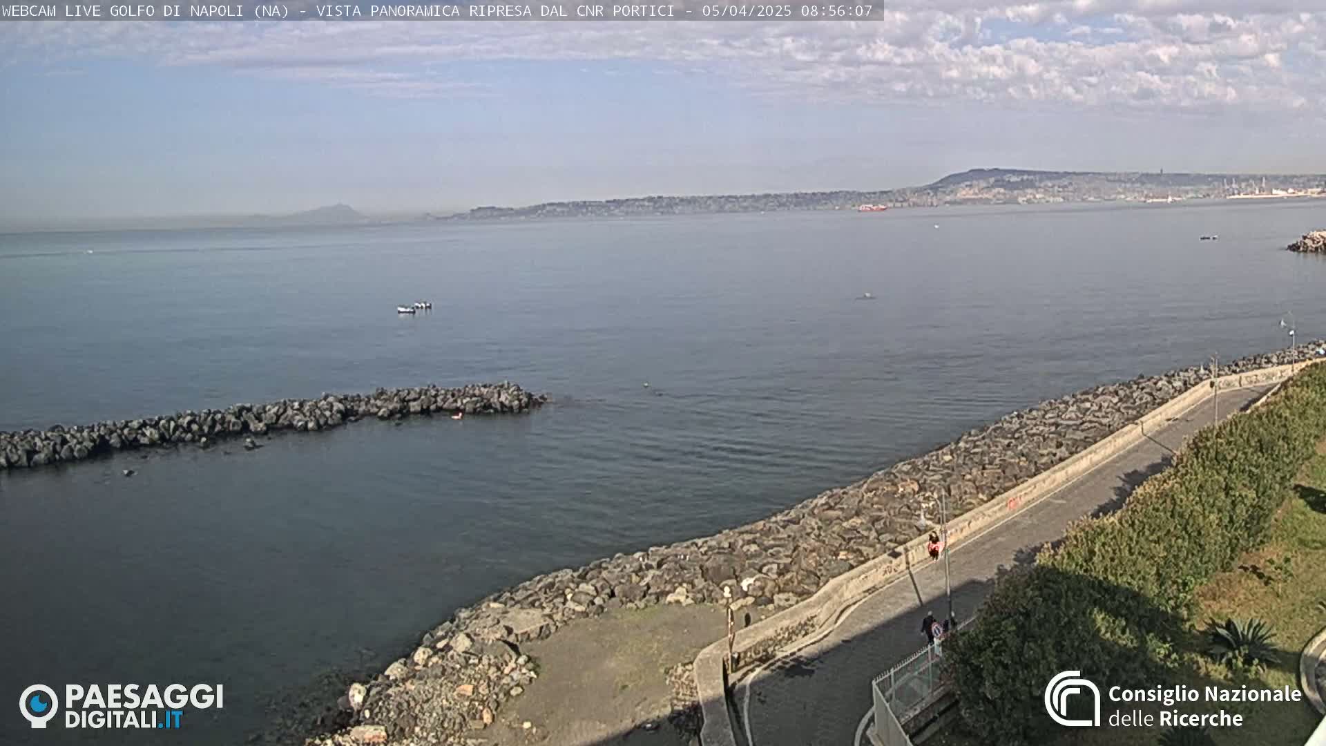 A rocky shoreline with a paved path and some greenery overlooks a calm sea under a partly cloudy sky, with a distant city and mountain visible on the horizon.