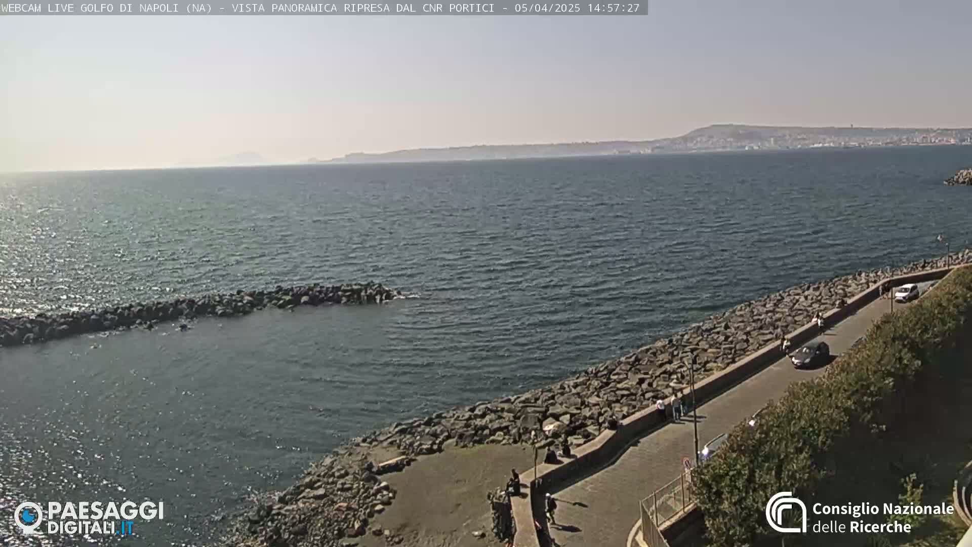 A calm sea with a rocky shoreline and a paved road, alongside which several people and cars are visible, under a mostly clear sky.