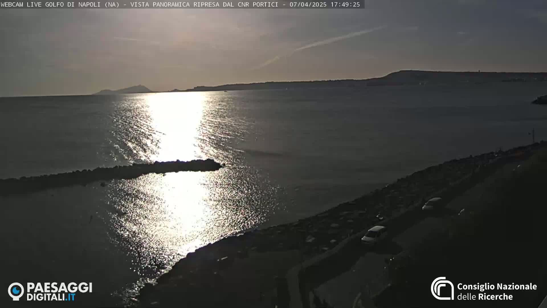 A calm sea reflects the setting sun, with a rocky shoreline and a few cars parked nearby.