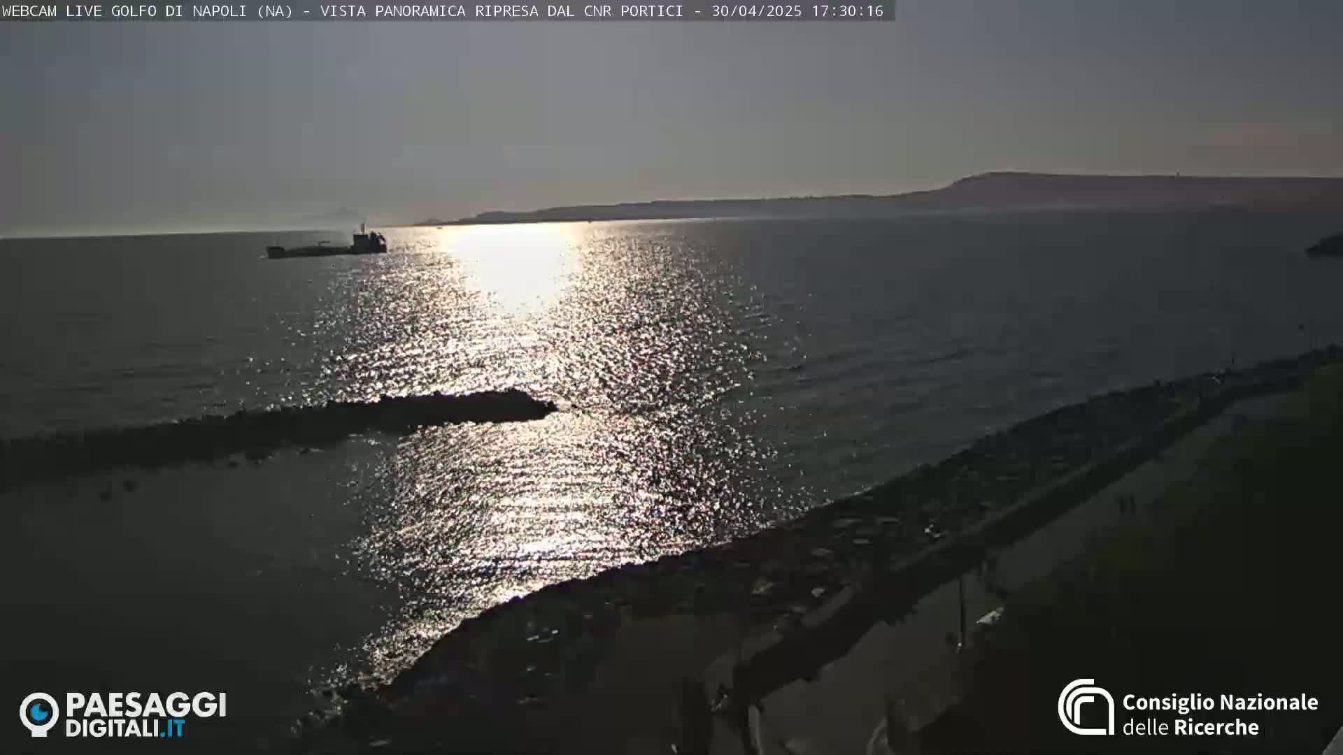 A cargo ship is visible on a calm, sun-lit sea near a rocky shore at sunset.