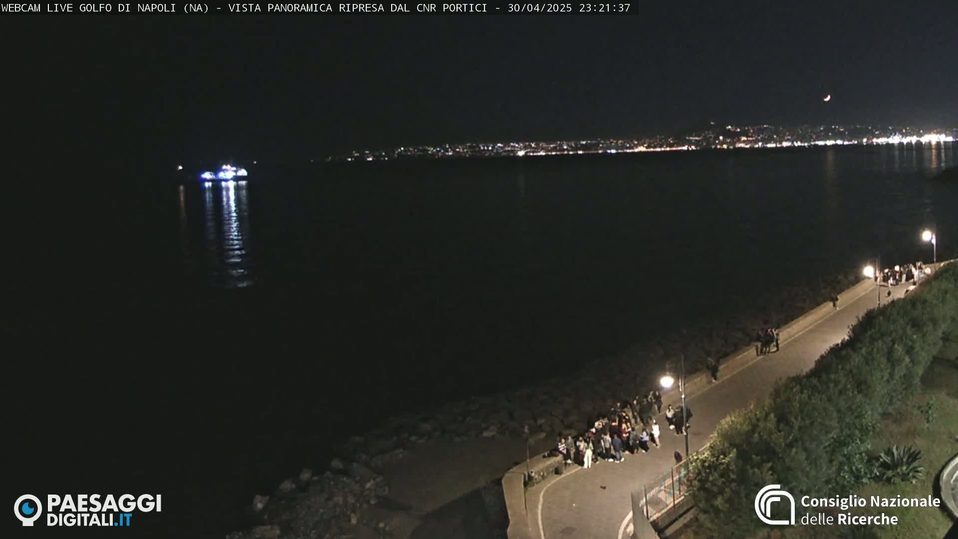 A nighttime view of a calm body of water with a distant city skyline, a boat on the water, and a group of people gathered on a paved walkway next to the water.
