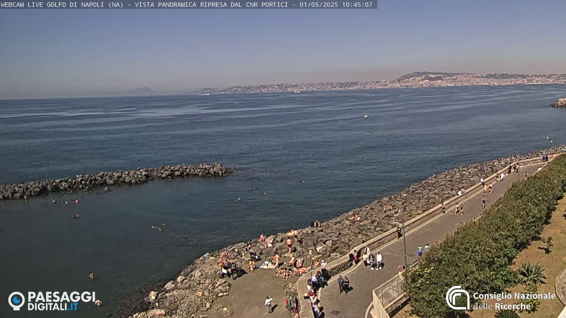 A rocky shoreline with sunbathers and people walking along a paved path, overlooks a calm blue sea under a clear sky, with a distant cityscape visible on the horizon.