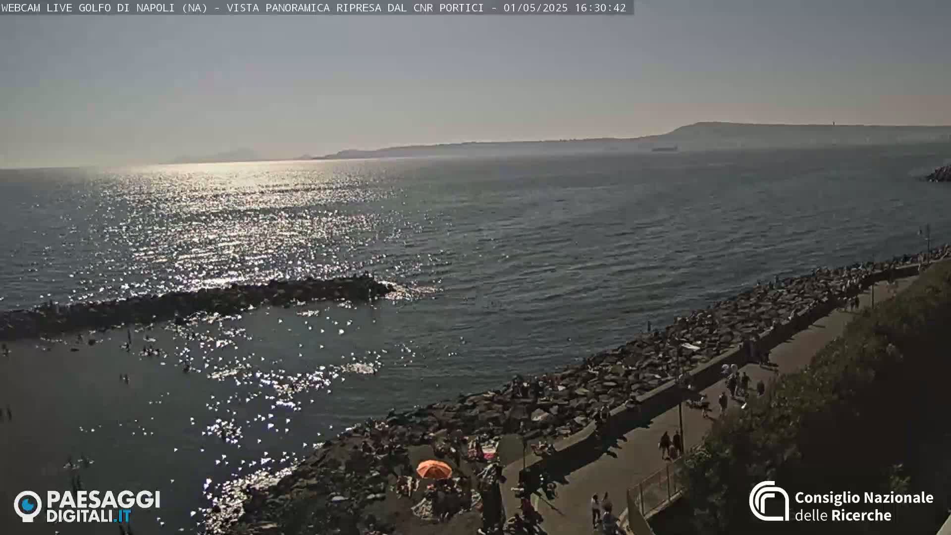 A sunny day at a rocky shoreline with people gathered on a path and in the water, a calm sea stretching to a distant landmass under a clear sky.