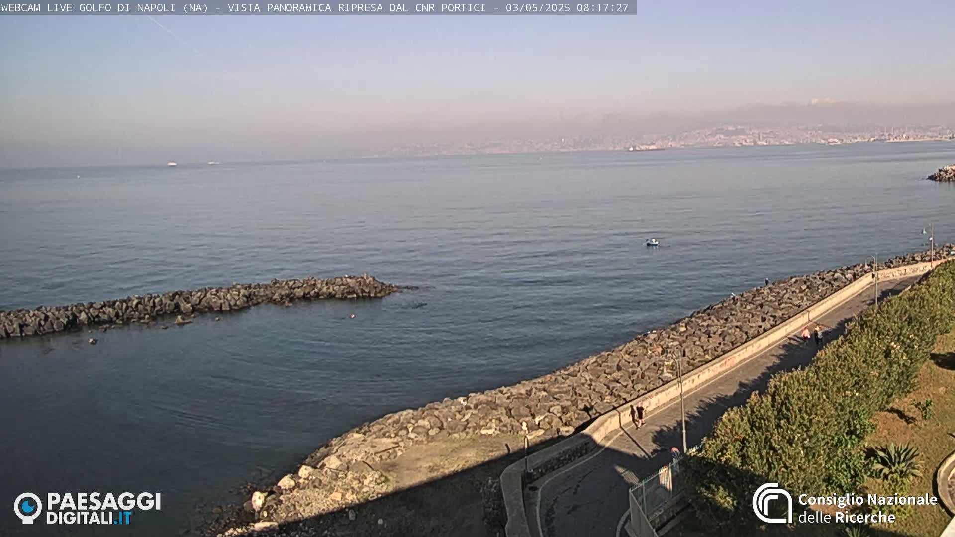 A hazy, sunny day reveals a rocky shoreline with a paved walkway and sparse pedestrians, overlooking a calm body of water extending to a distant city and mountains.