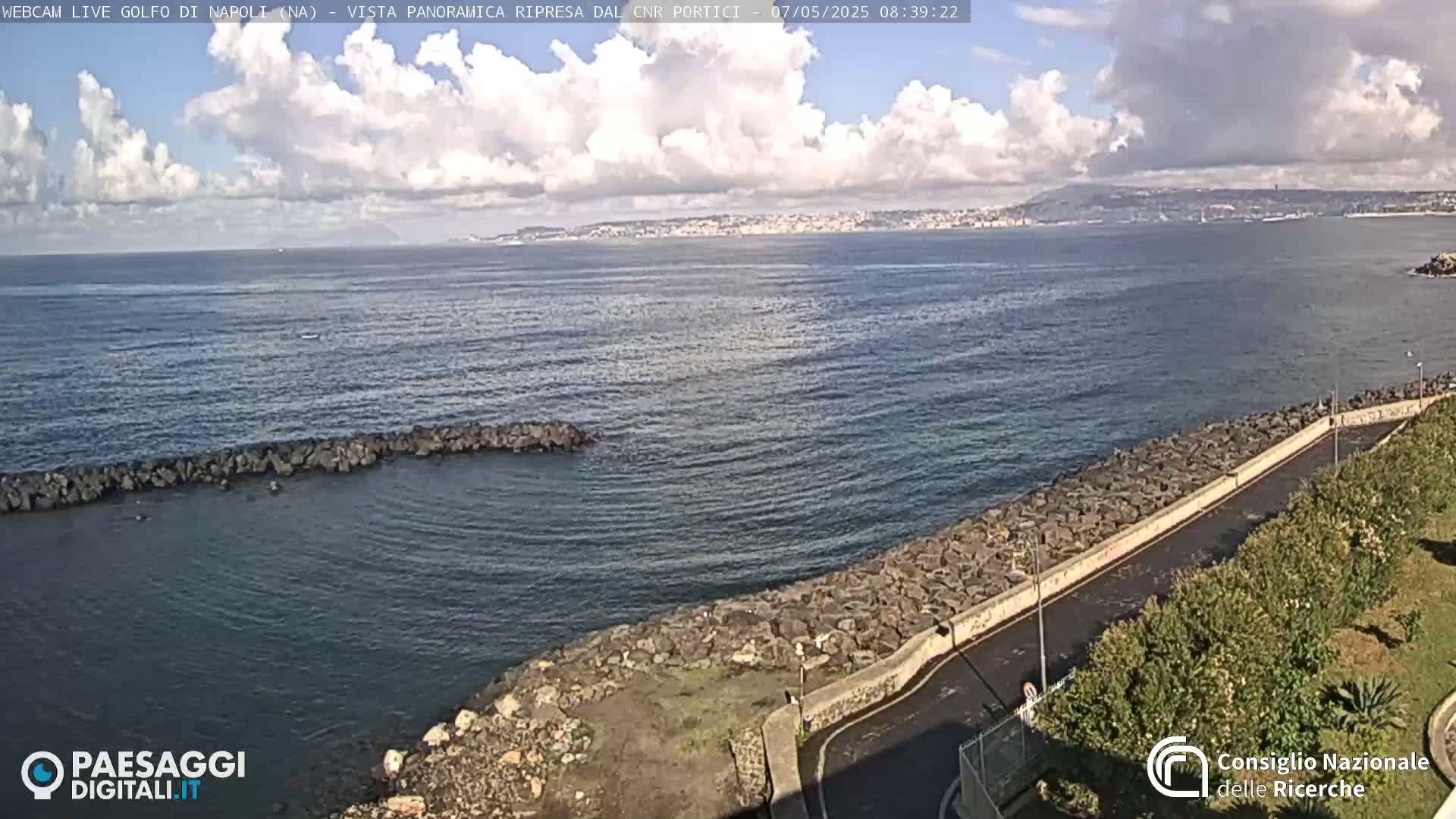 A partly cloudy day shows a rocky shoreline and calm sea with a distant city visible across the water.